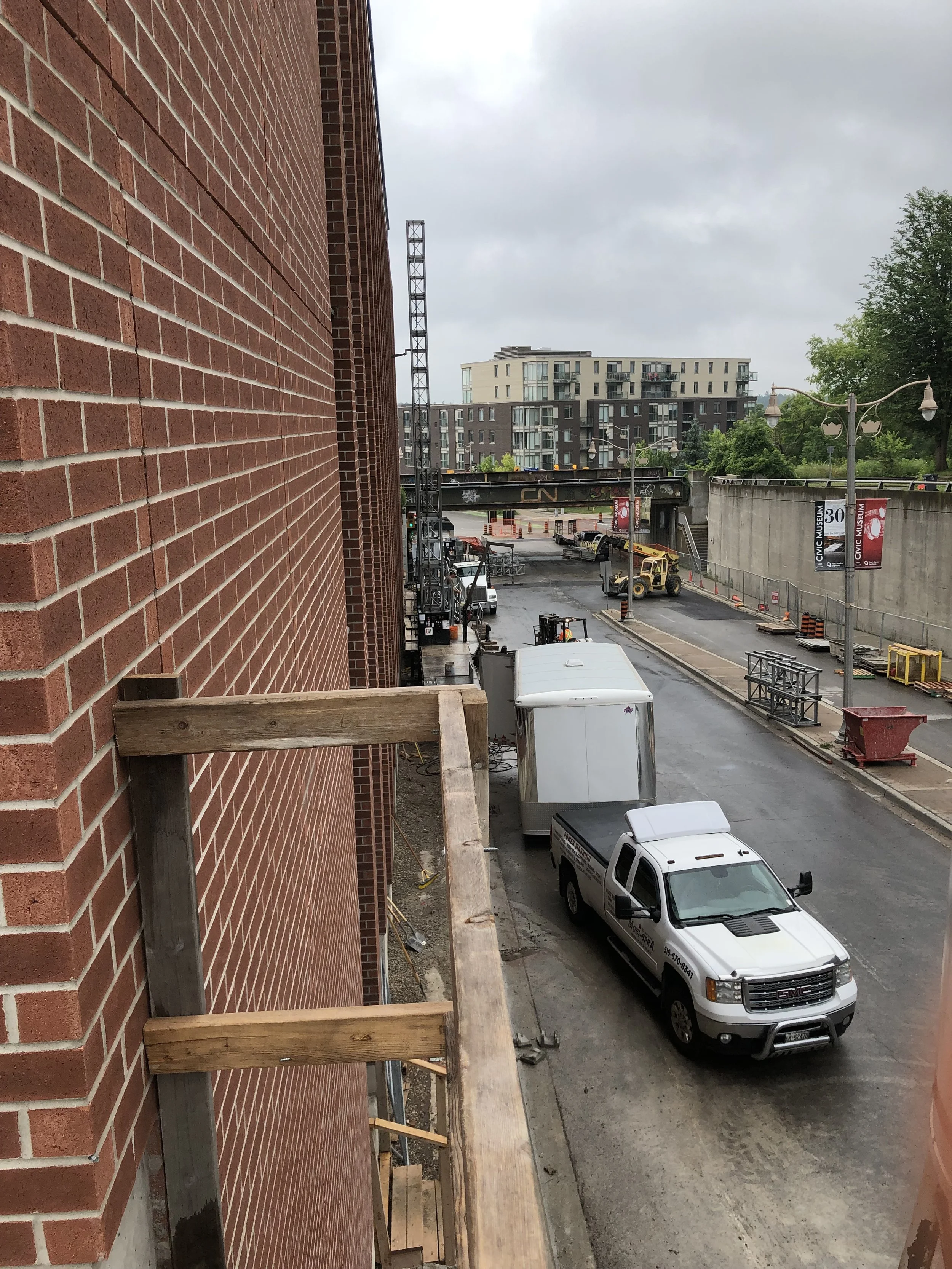 A view from a balcony shows a construction area with vehicles, equipment, and scaffolding adjacent to a brick building. In the background, there are modern apartment buildings and a bridge overpass.