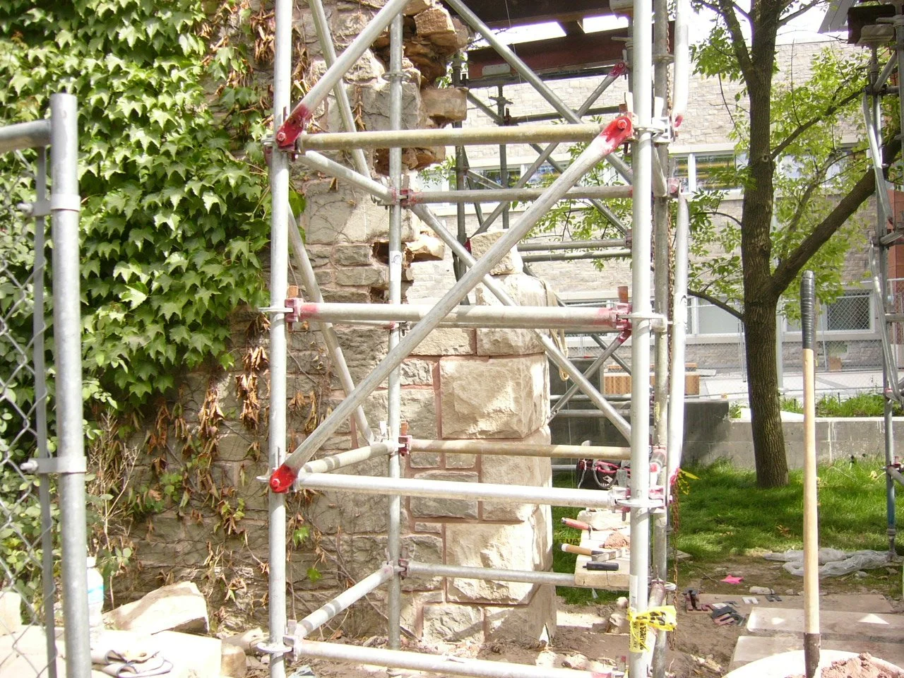 A construction site with metal scaffolding around a stone wall with wooden beams, a tree, and green foliage.