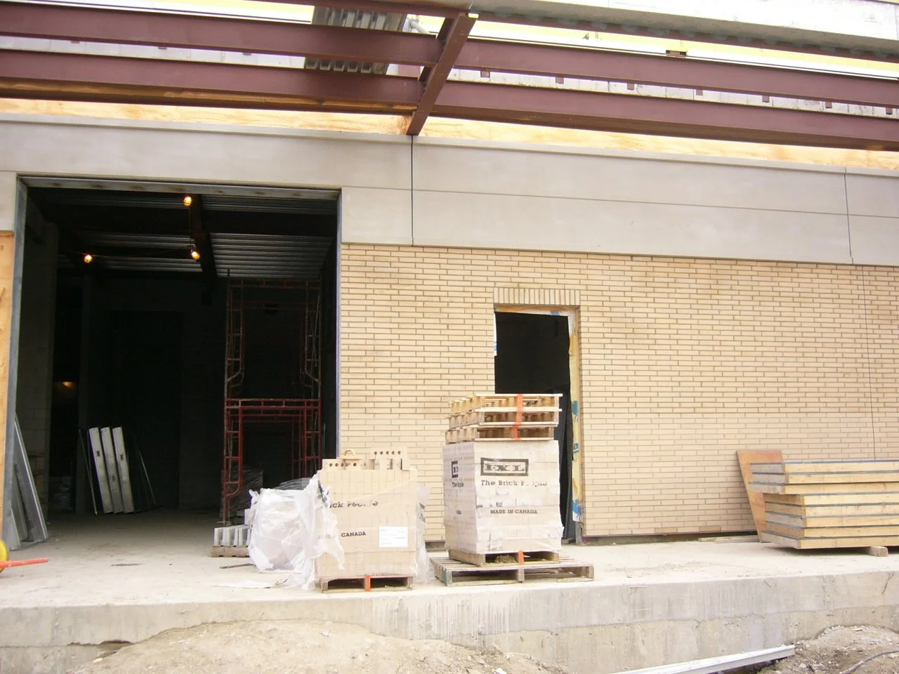 Construction site showing a partly built brick wall with a door opening, stacks of construction materials, and an open garage or storage area under construction.