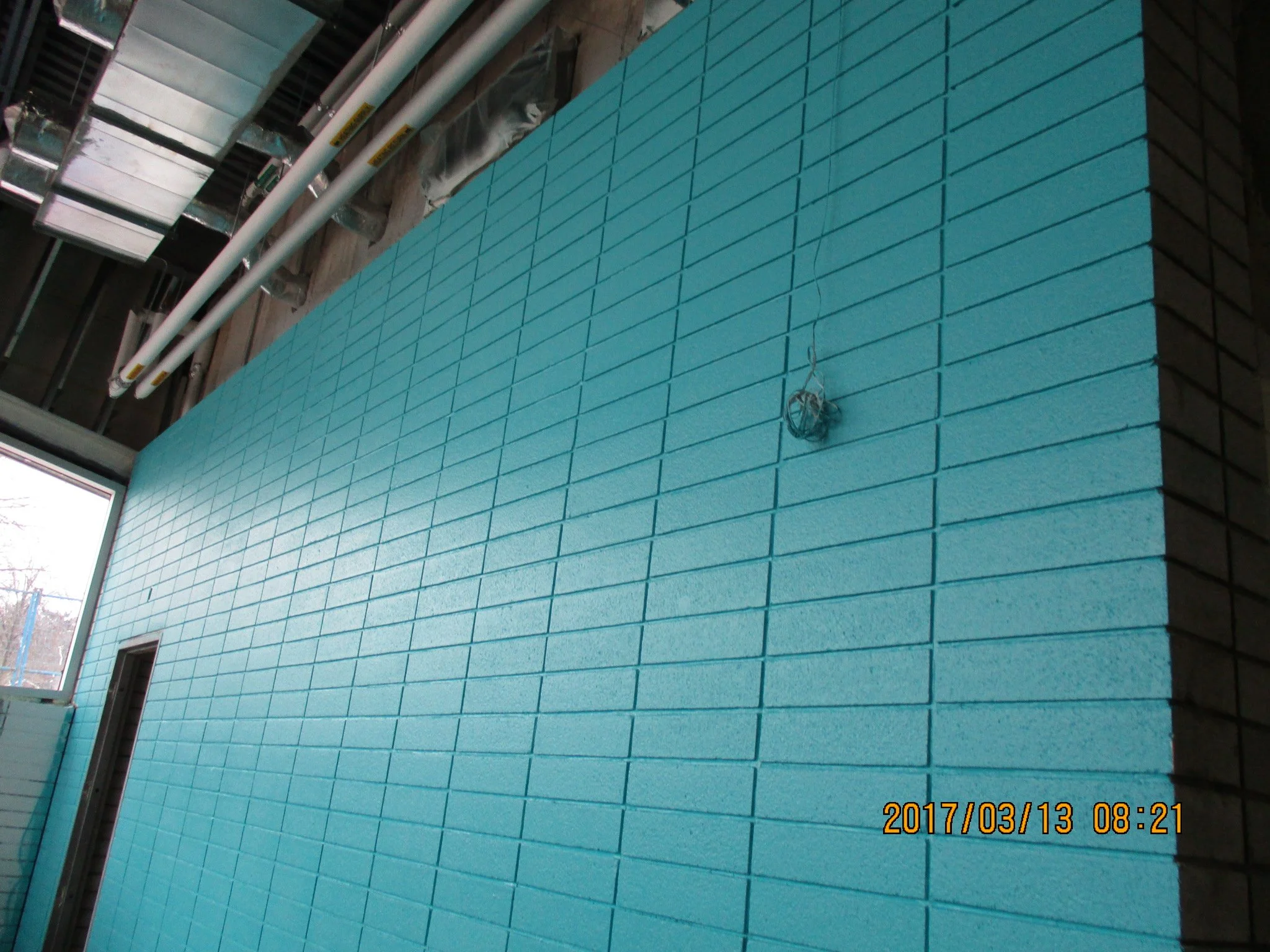 Interior of a building under construction with a large wall painted turquoise, brick pattern, with visible pipes and conduit, and a small window on the left.