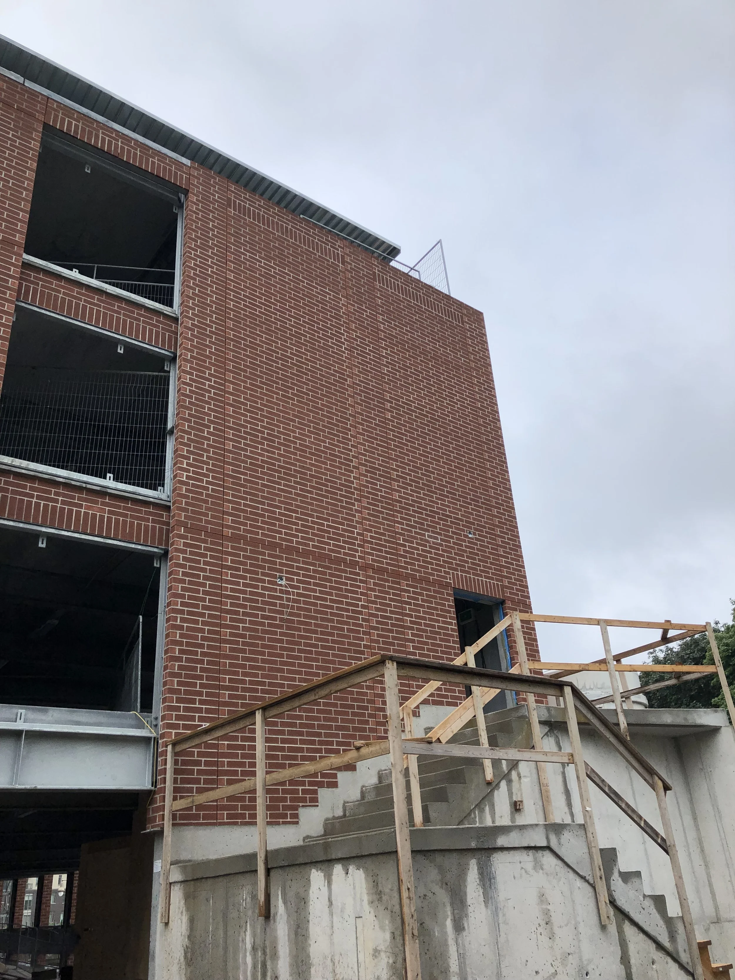Under construction brick building with stairs and wooden handrails leading to a door, with open spaces for future windows.