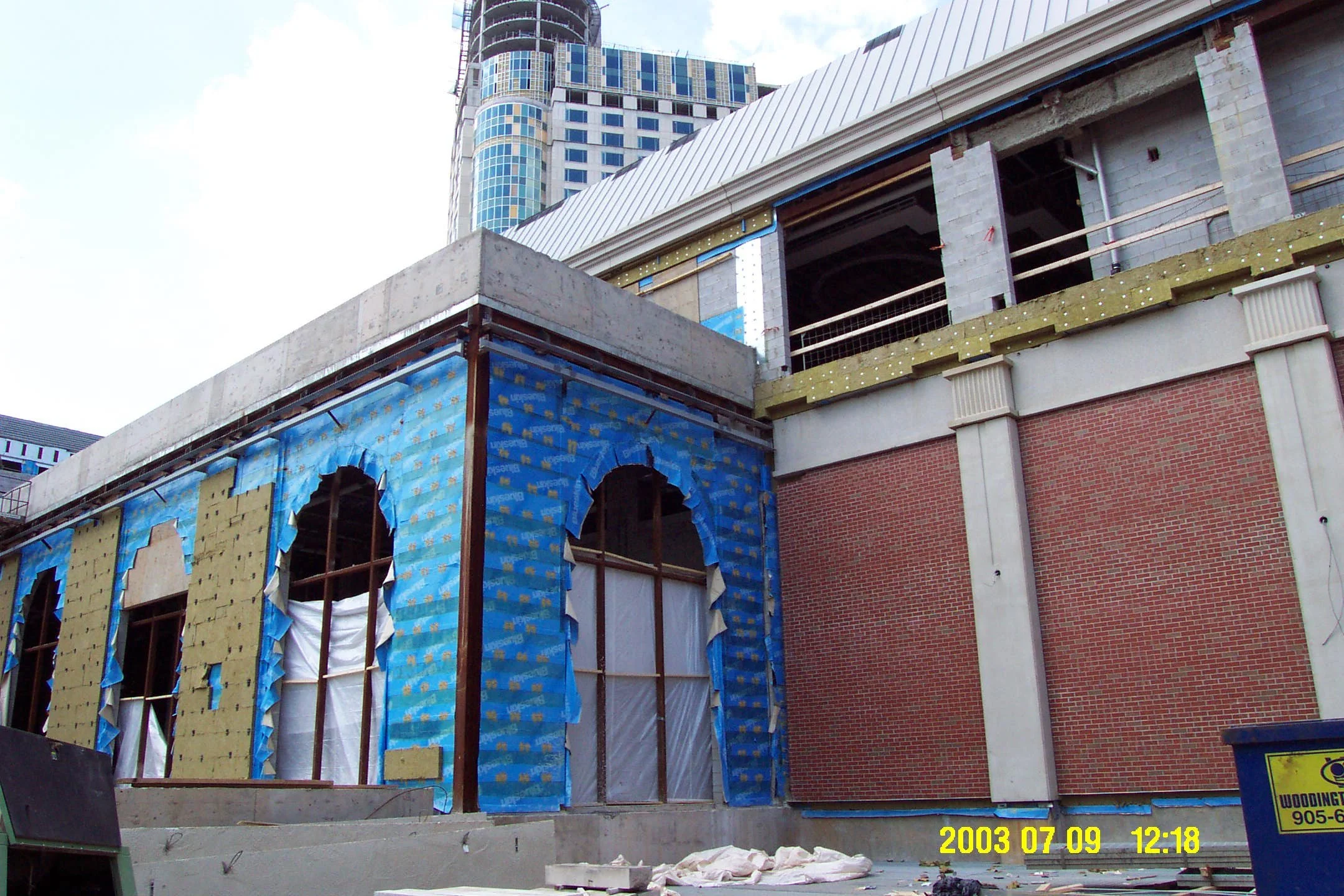 Construction site with building in progress, showing brick and concrete walls, blue insulation wrap, and window frames, with a tall modern skyscraper in the background, under a mostly cloudy sky.