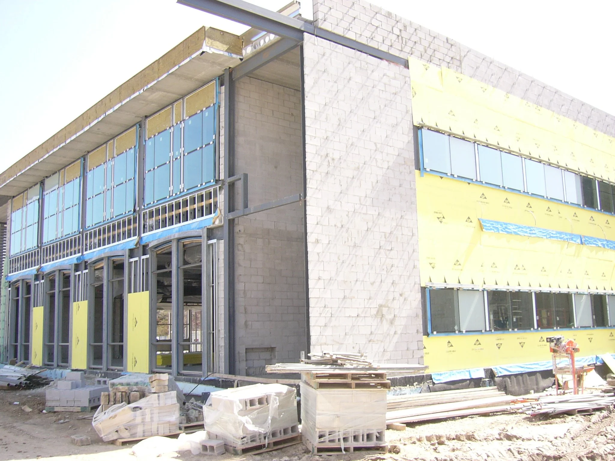 Under construction building with yellow insulation, metal framing, and large windows, construction materials on the ground.