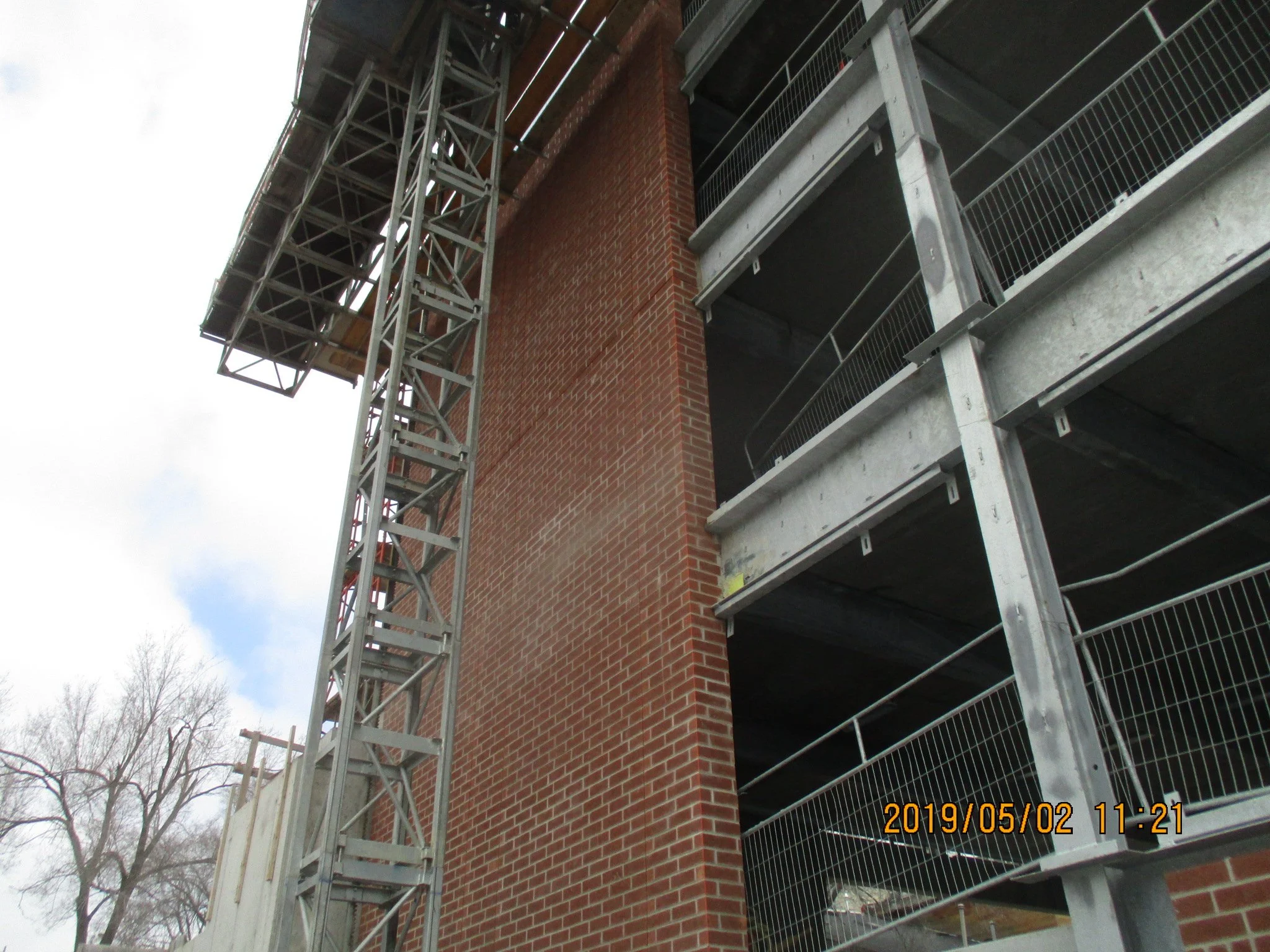 Construction site with a brick building and an external metal staircase. A construction crane or tower lift is also visible, with part of the sky and trees in the background.
