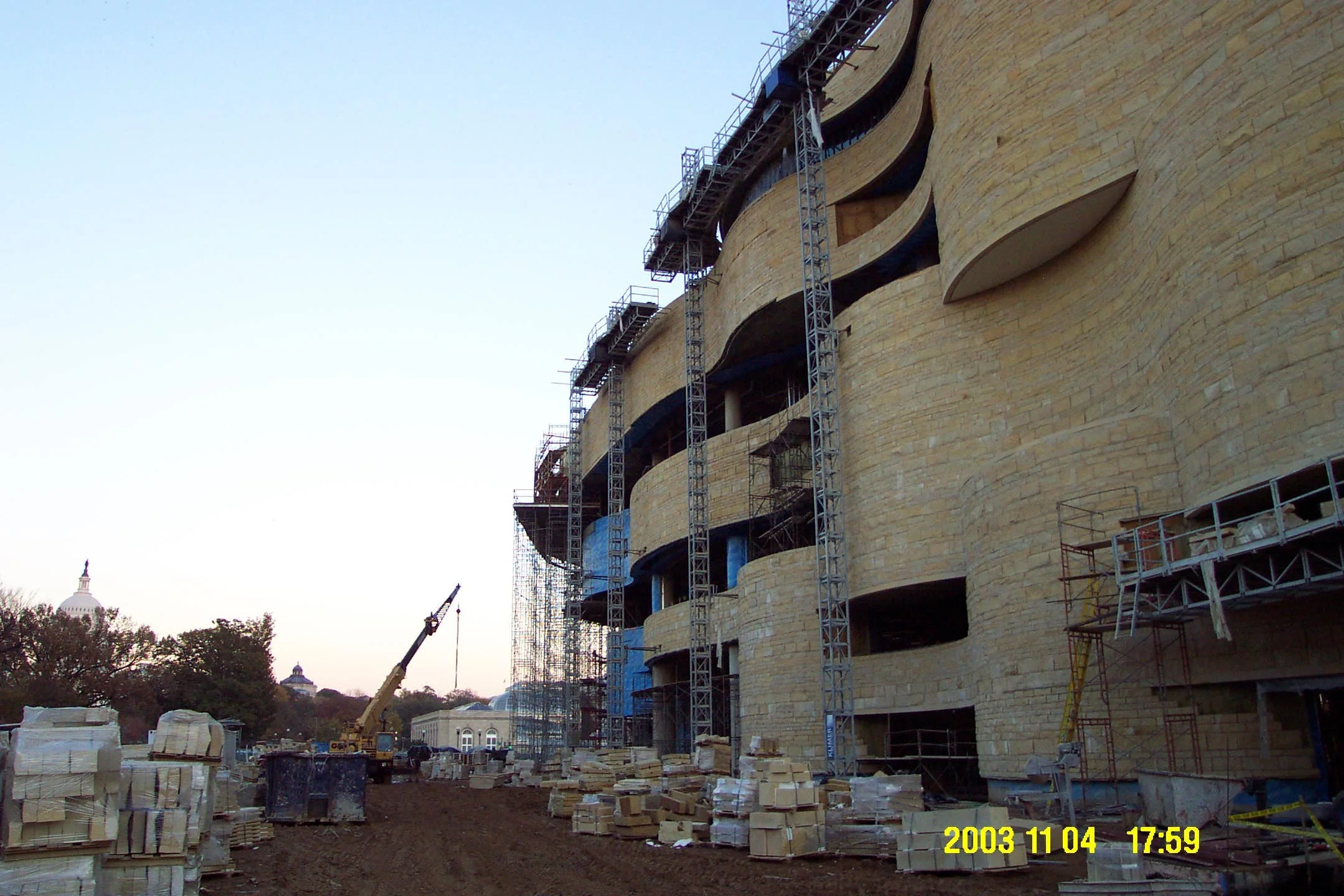 Construction site of a building with scaffolding, stone facade, and construction materials, with a crane and distant dome-shaped structures in the background, taken in early evening.
