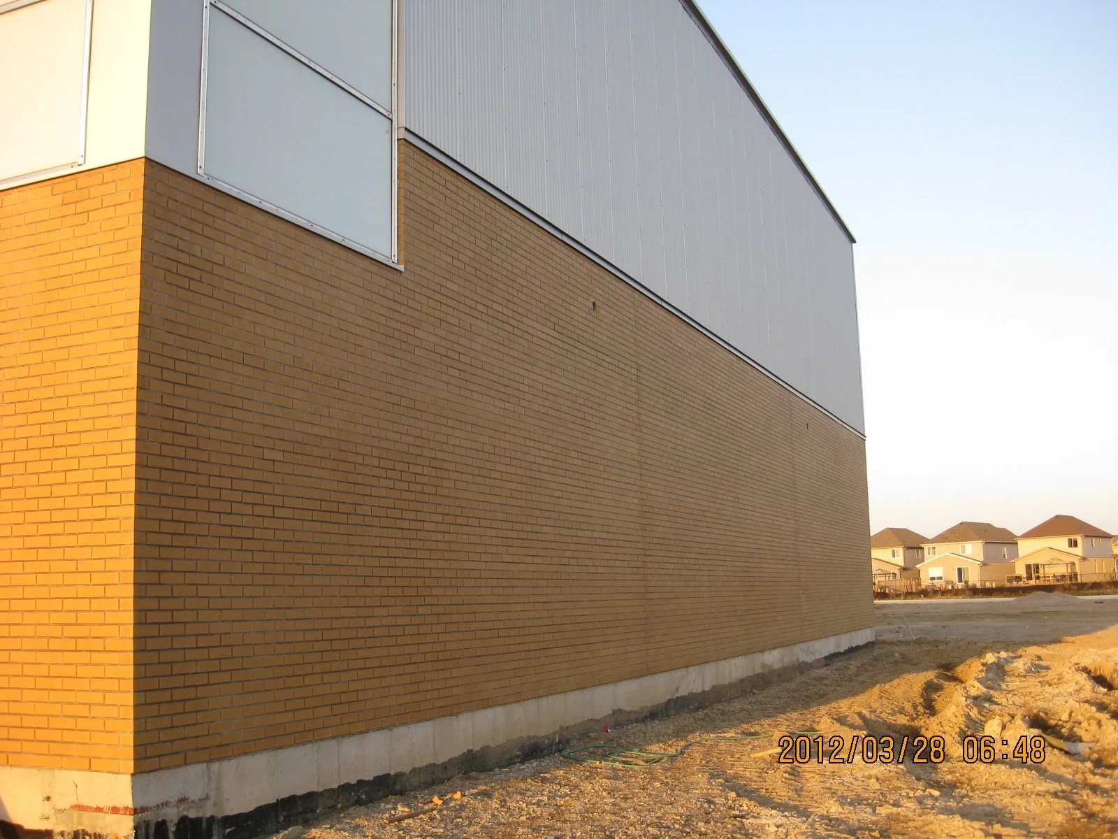 Side view of a building with a brick wall and metal siding on the upper part, with a row of houses in the distant background and a dirt ground in the foreground.