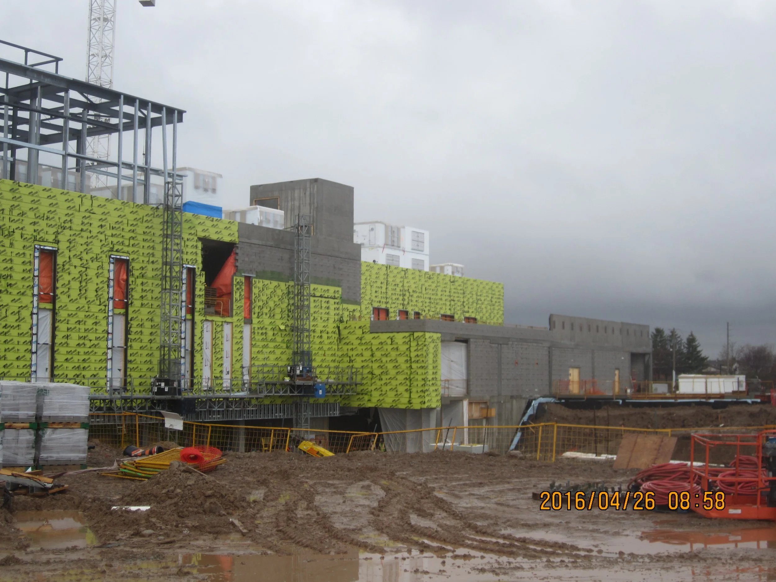 Construction site with partly built green insulations on building walls, dirt ground, construction equipment, and cloudy sky.