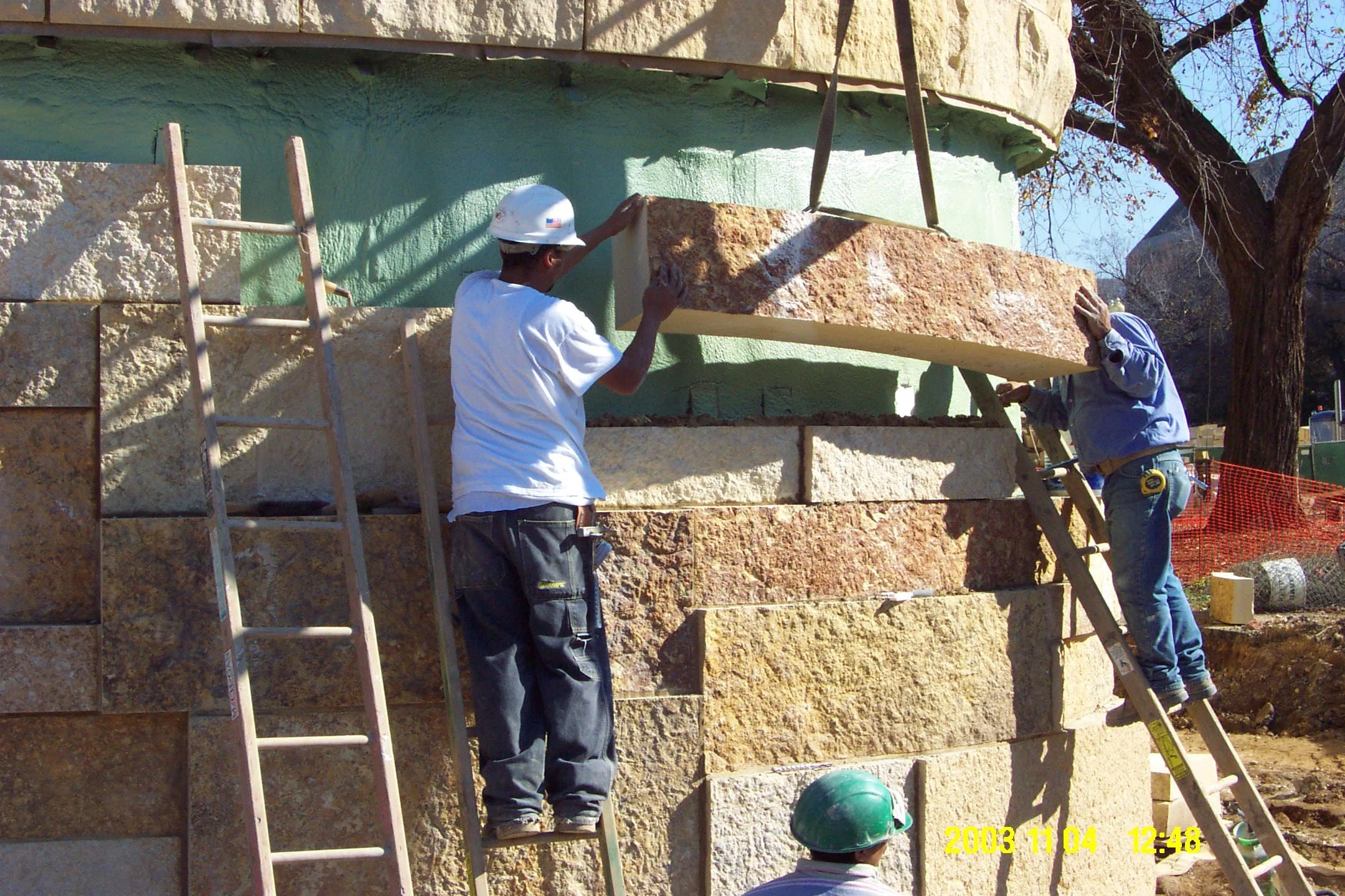 Construction workers installing large stone blocks on the exterior of a building, using ladders and safety equipment, with trees and orange safety fencing in the background.