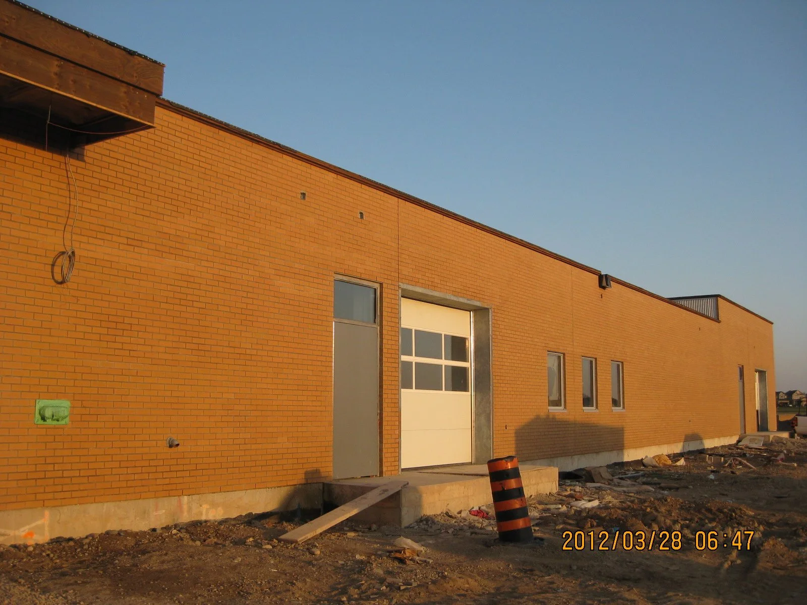 A building under construction with a brick exterior, a white garage door, and small windows, set against a clear blue sky, with construction materials and debris on the ground.