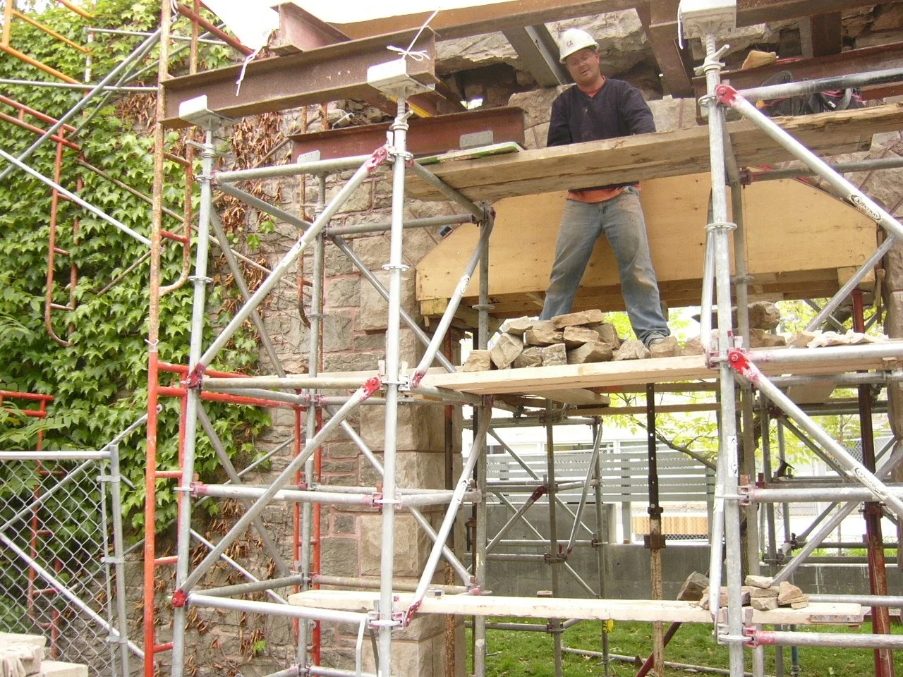 A construction worker wearing a white safety helmet and dark clothing stands on a wooden platform among metal scaffolding, working on a stone wall with rocks and a wooden support structure.