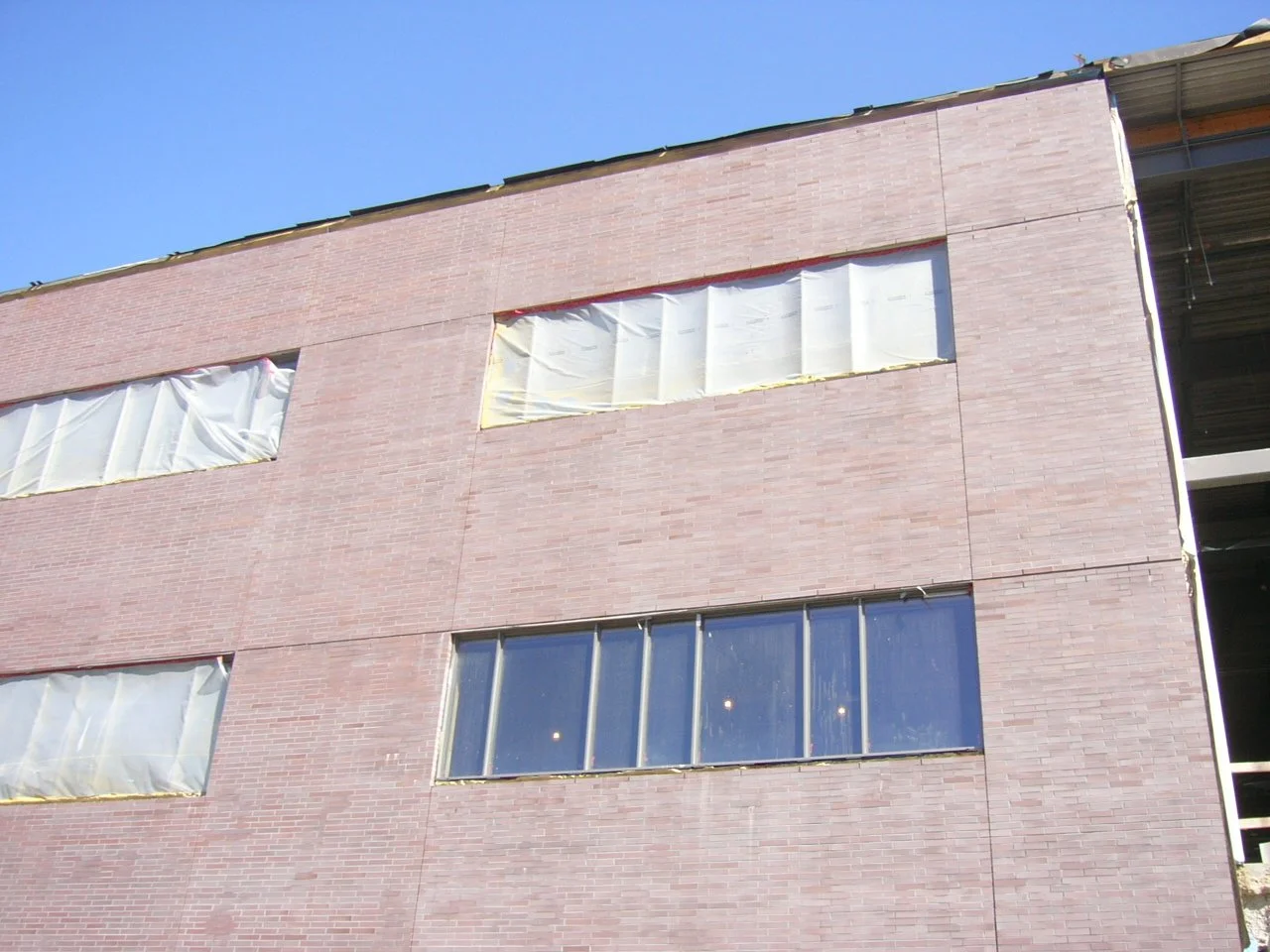 A multi-story building under construction or renovation with a brick exterior, showing three rectangular windows and covered openings wrapped in plastic, against a clear blue sky.