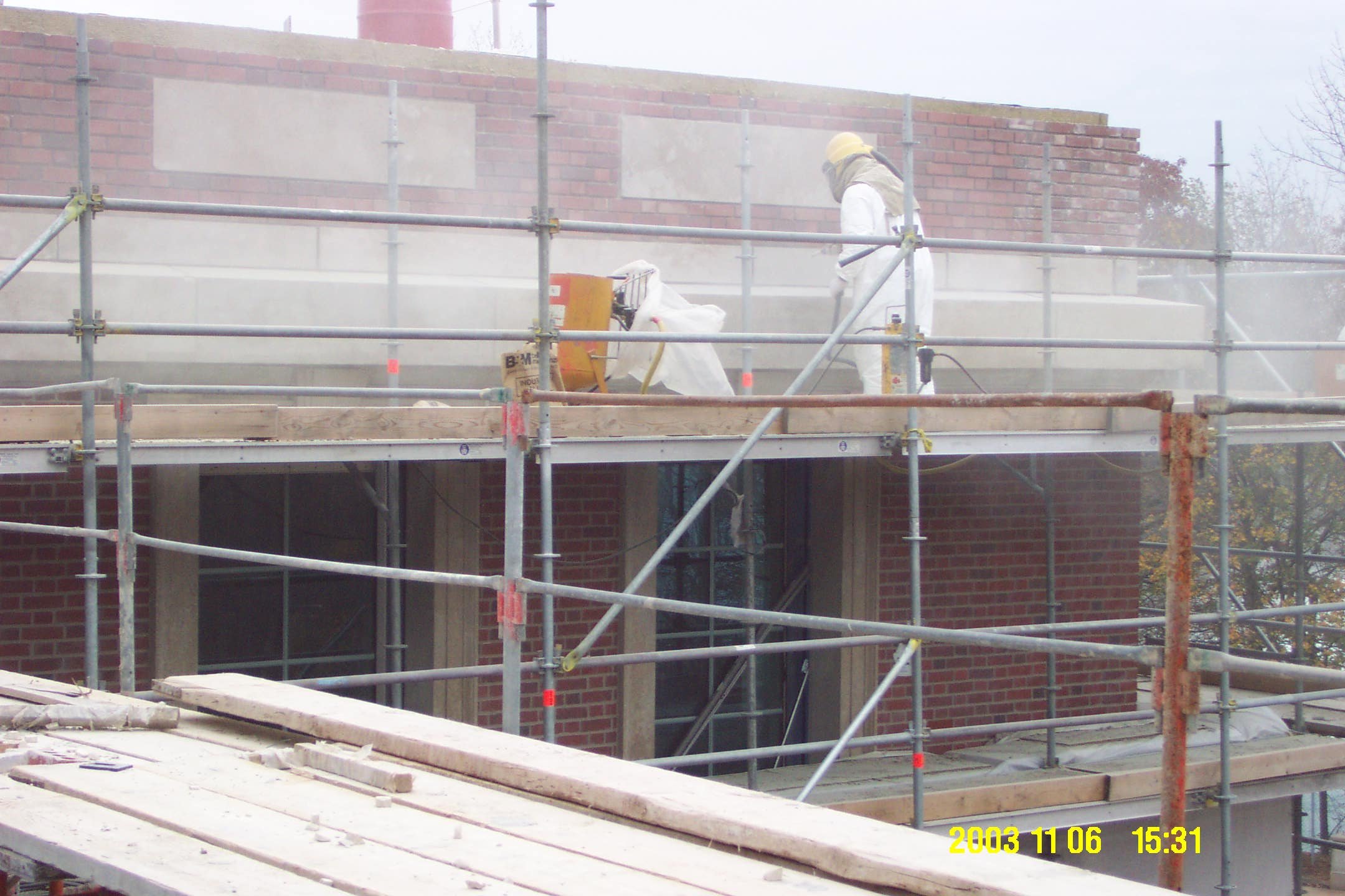 Construction worker in white protective suit and yellow hard hat working on scaffolding outside a brick building, with foggy atmosphere and trees in the background.