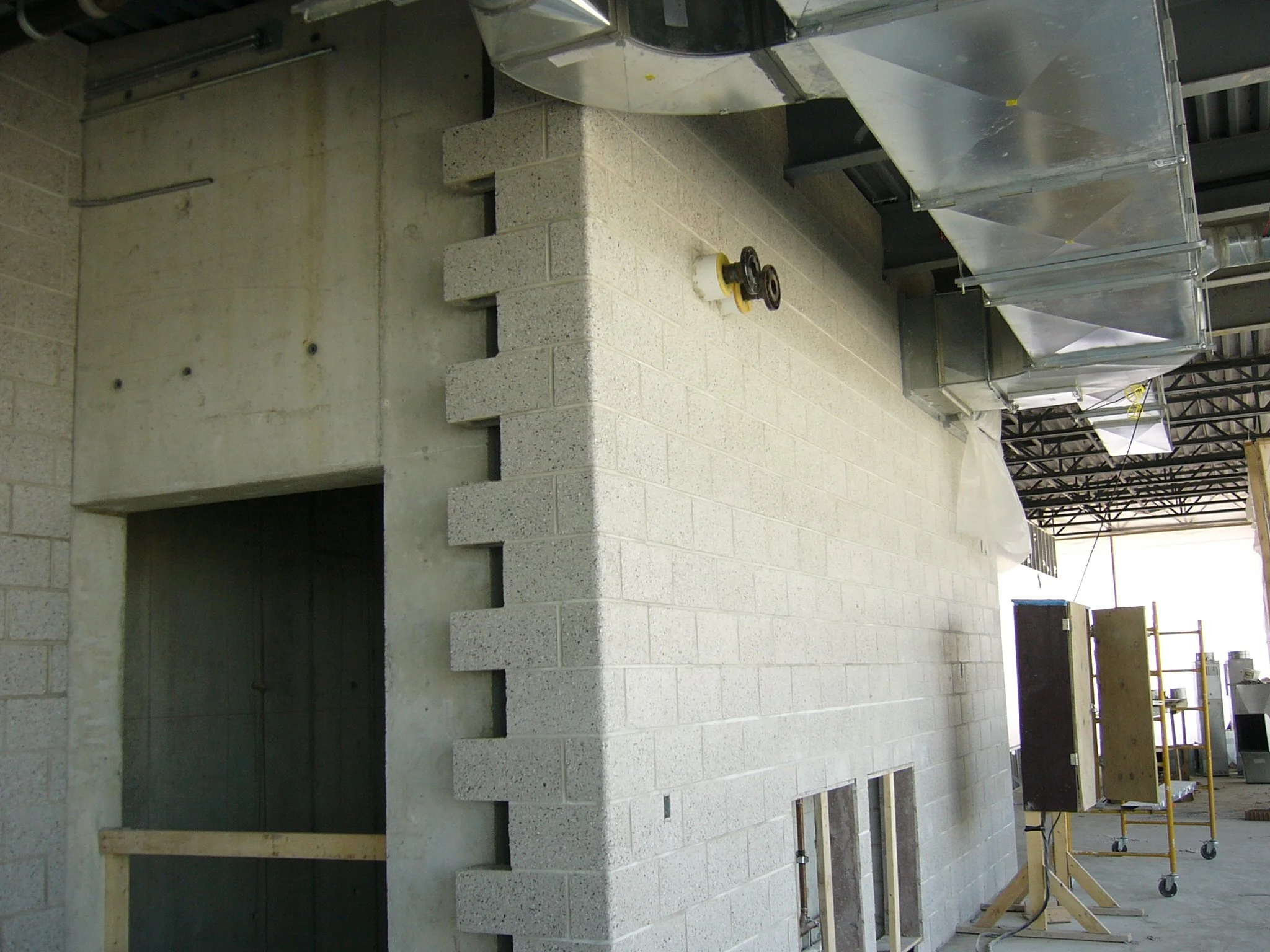 Construction site showing a partially built wall with concrete blocks, construction tools, and ventilation ductwork in an indoor environment.