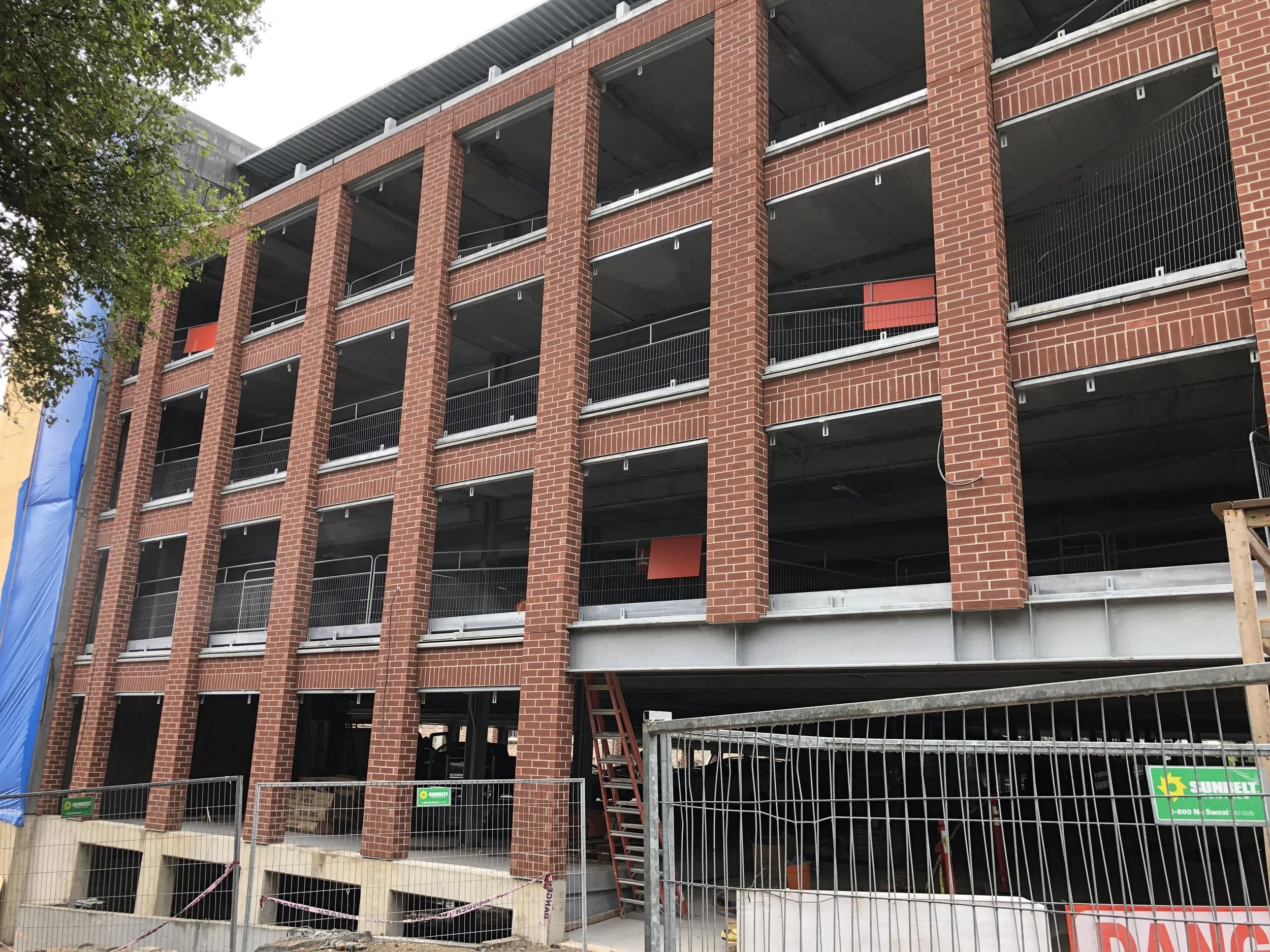 Under construction multi-story brick building with open parking garage levels, construction fencing, and safety signage.