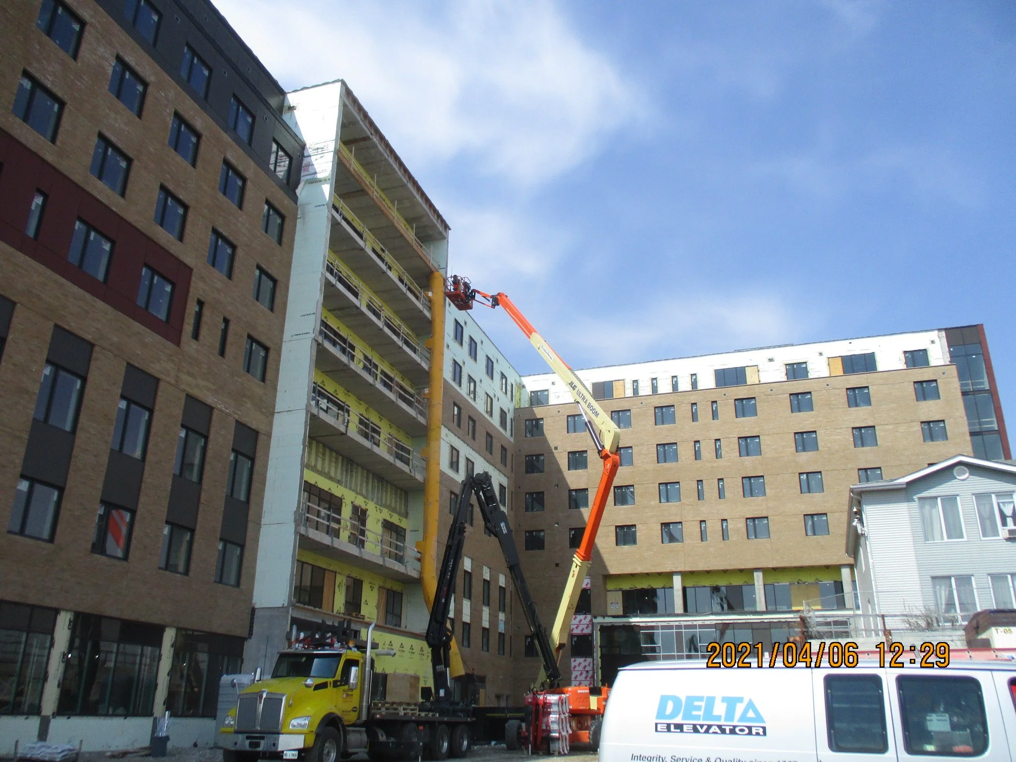 Construction site with a large building in the process of being built. An orange boom lift is extending to the upper floors of the building, with workers on the platform. A yellow truck with a black arm is parked nearby, and the scene is under a part