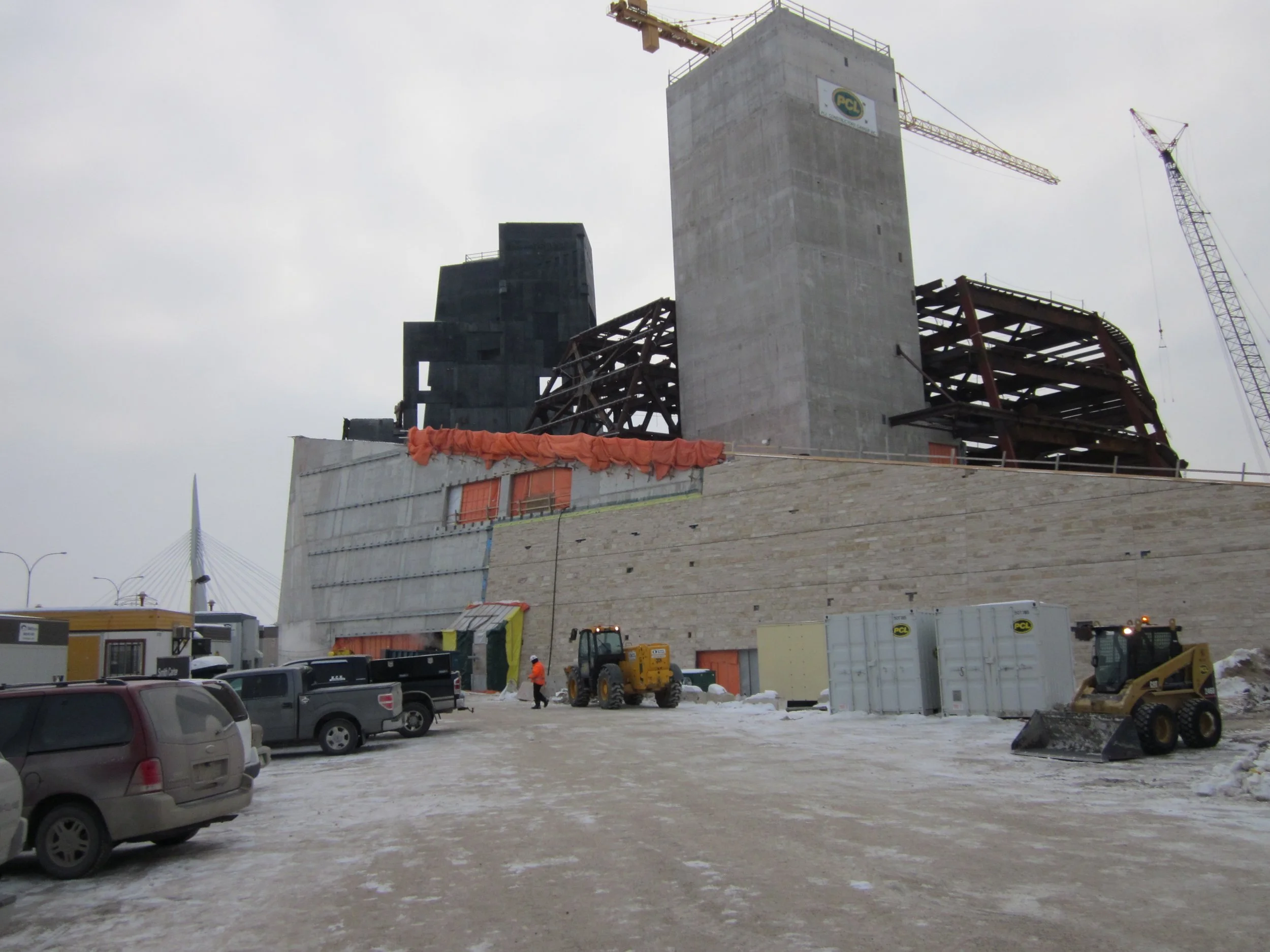 Construction site with unfinished building, cranes, vehicles, and workers in winter weather.
