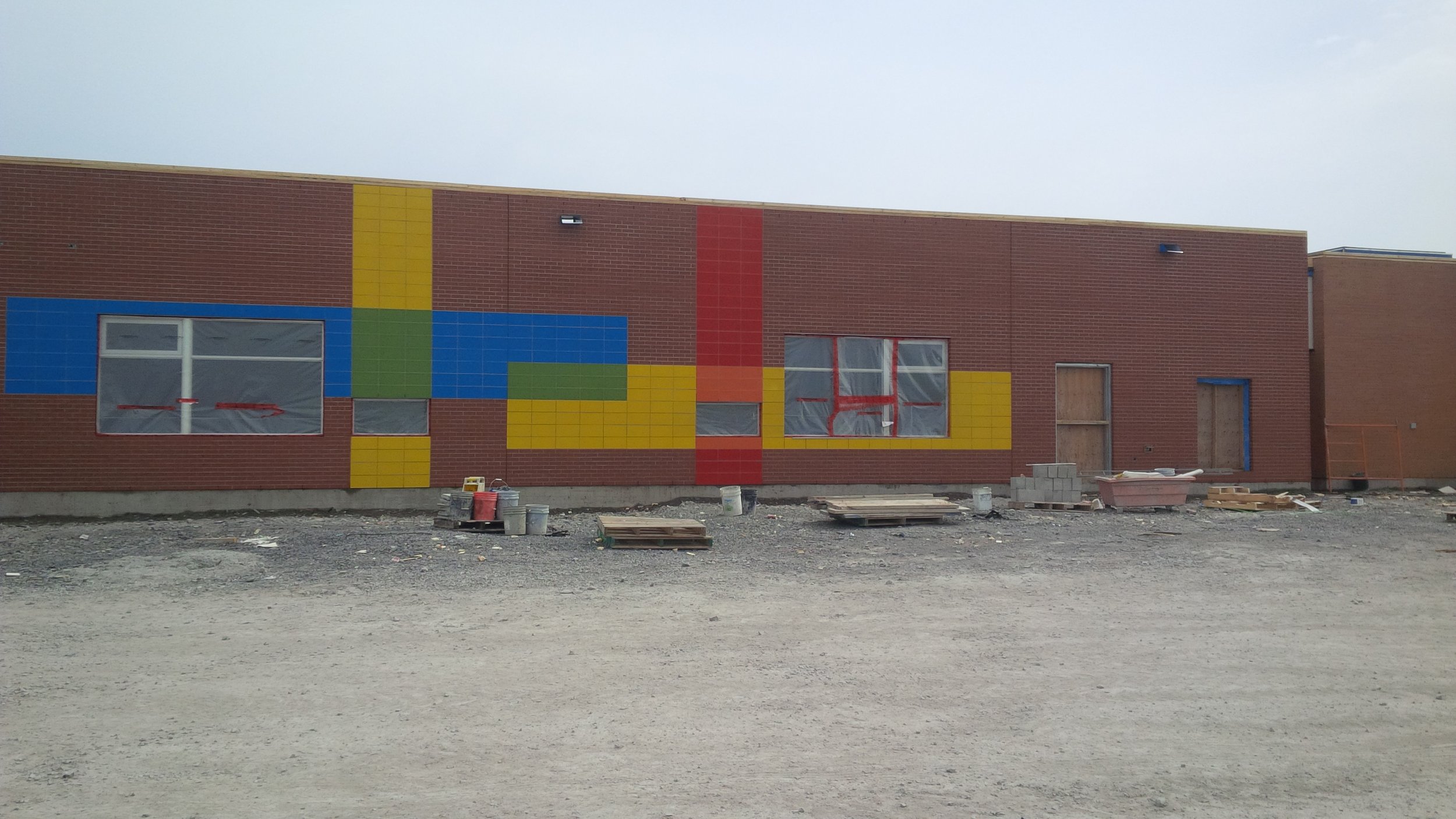 Building under construction with a brick exterior, featuring a colorful pixel art design in blue, green, yellow, red, and orange tiles on the wall. Windows are covered with plastic, and construction materials are in front.