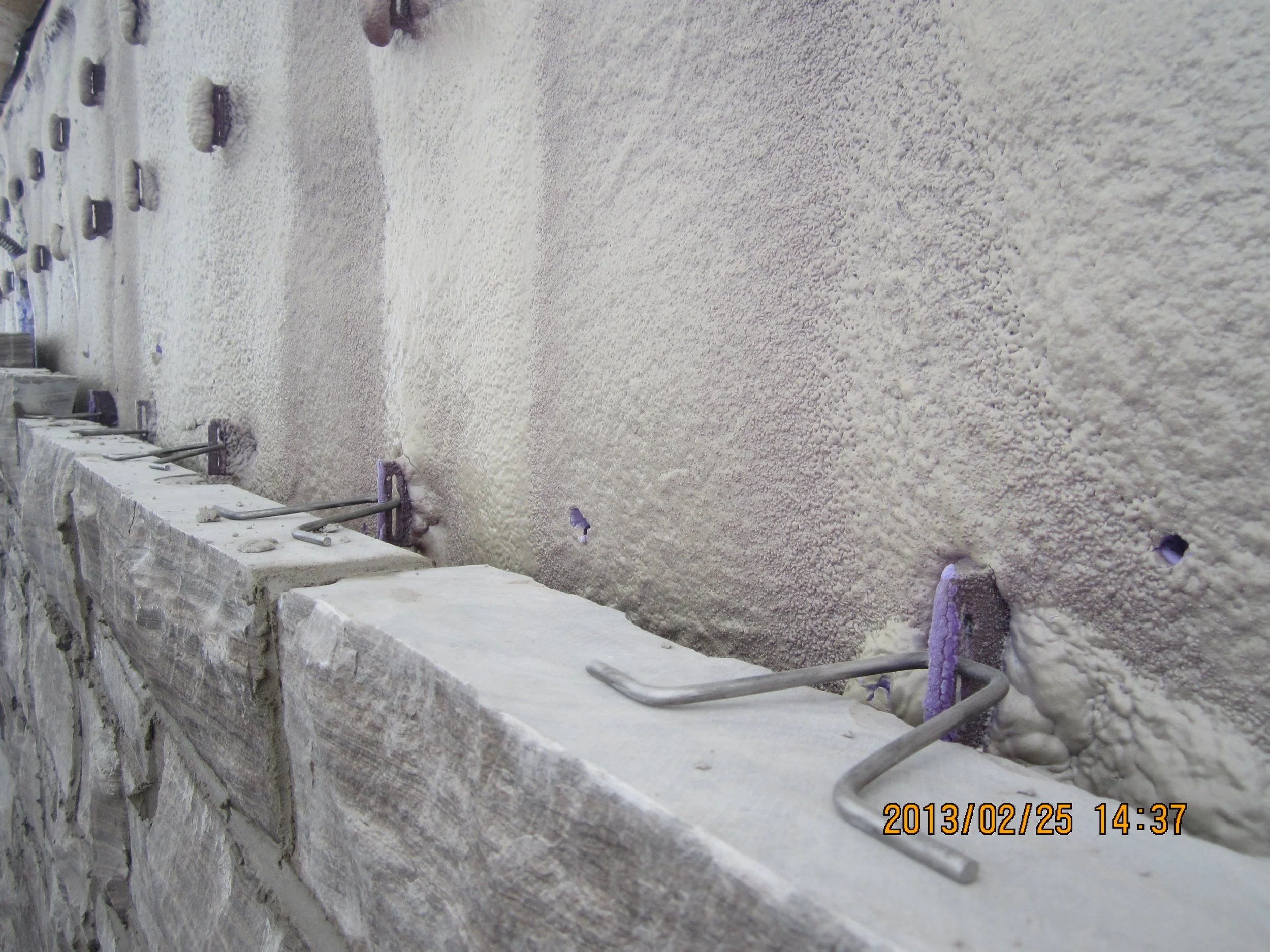 Close-up of a wall with spray foam insulation and metal support laths, with a stone ledge at the bottom.