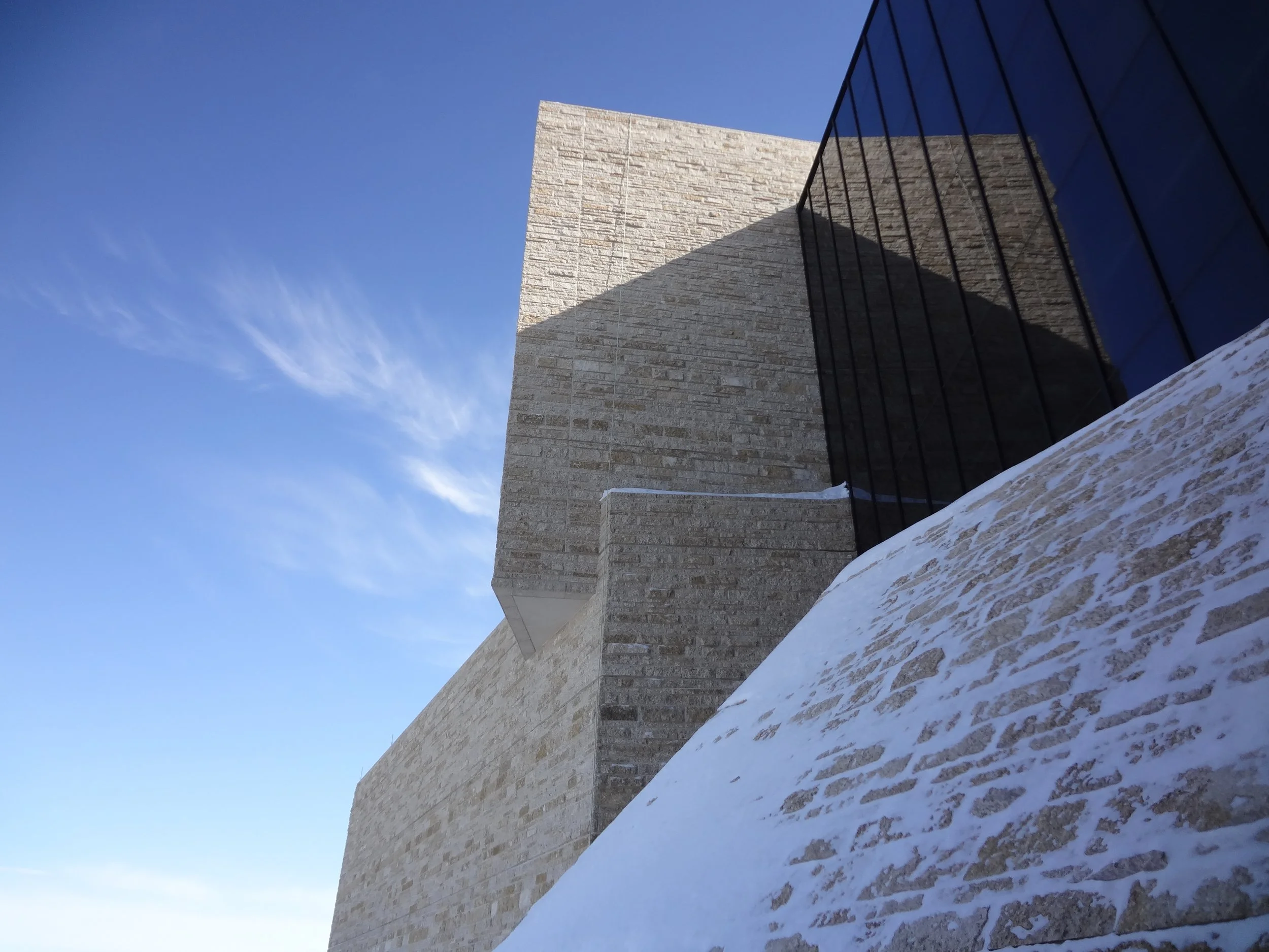 Low-angle shot of a modern building with beige brick walls, black glass windows, and snow on the ground against a clear blue sky.
