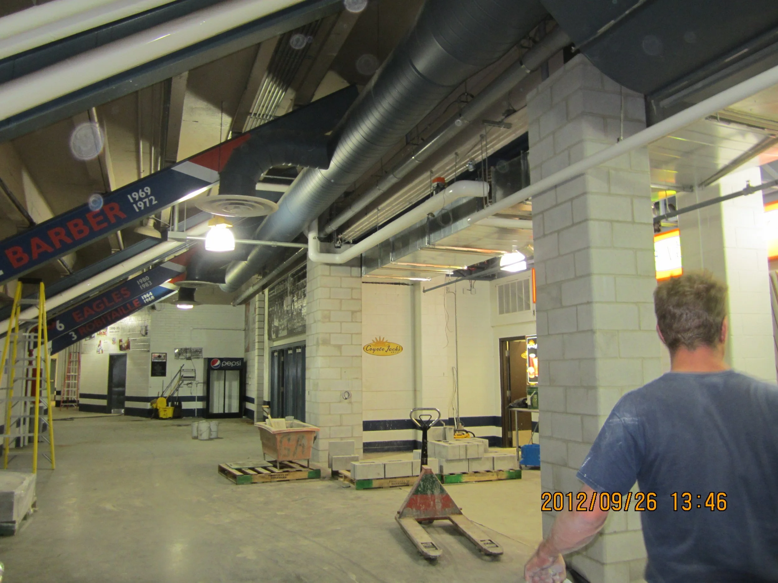 Interior of a construction site or sports facility with exposed ductwork, signage, and construction materials. A man with gray hair wearing a blue t-shirt stands with his back to the camera near a brick column.