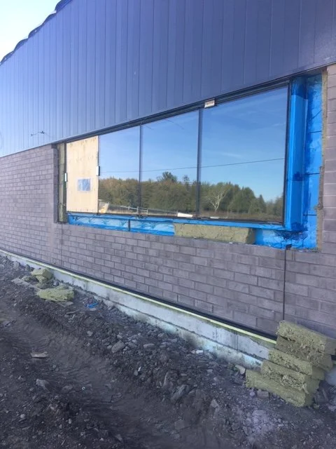 Building under construction with brick and siding exterior, large window installation underway, construction materials and dirt ground visible.
