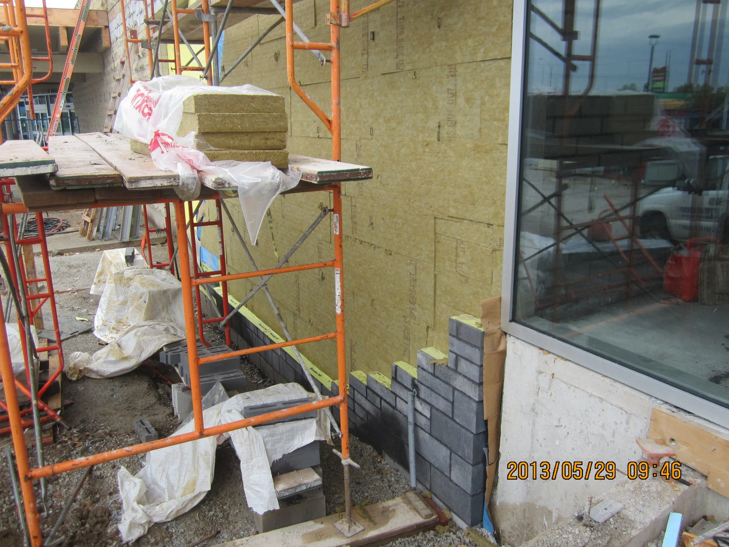 Construction site with Orange scaffolding, insulation panels, and a partially constructed brick wall next to a glass window.