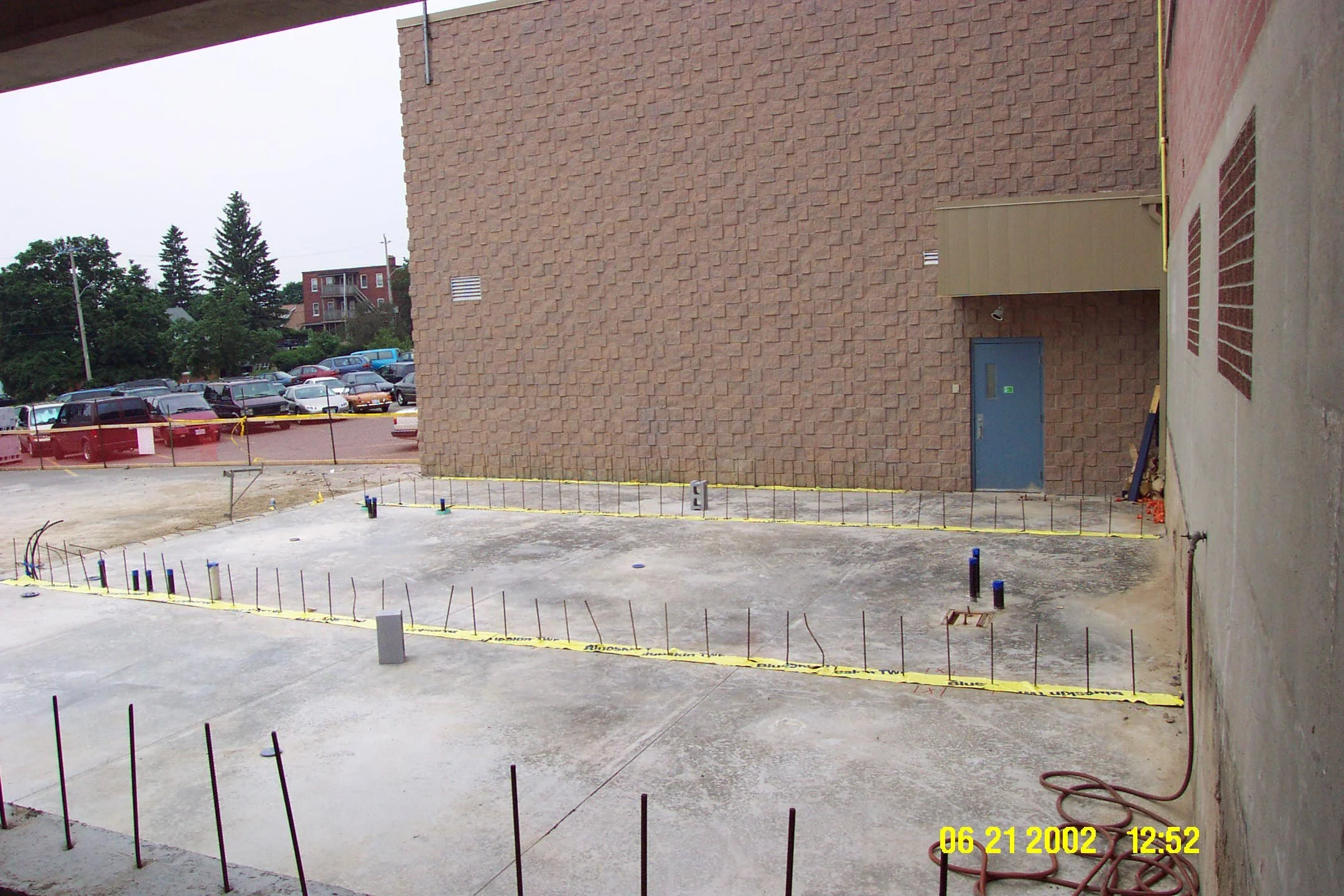 Construction site with a concrete slab, metal rods, and pipes, next to a brick building, with a parking lot and trees in the background.