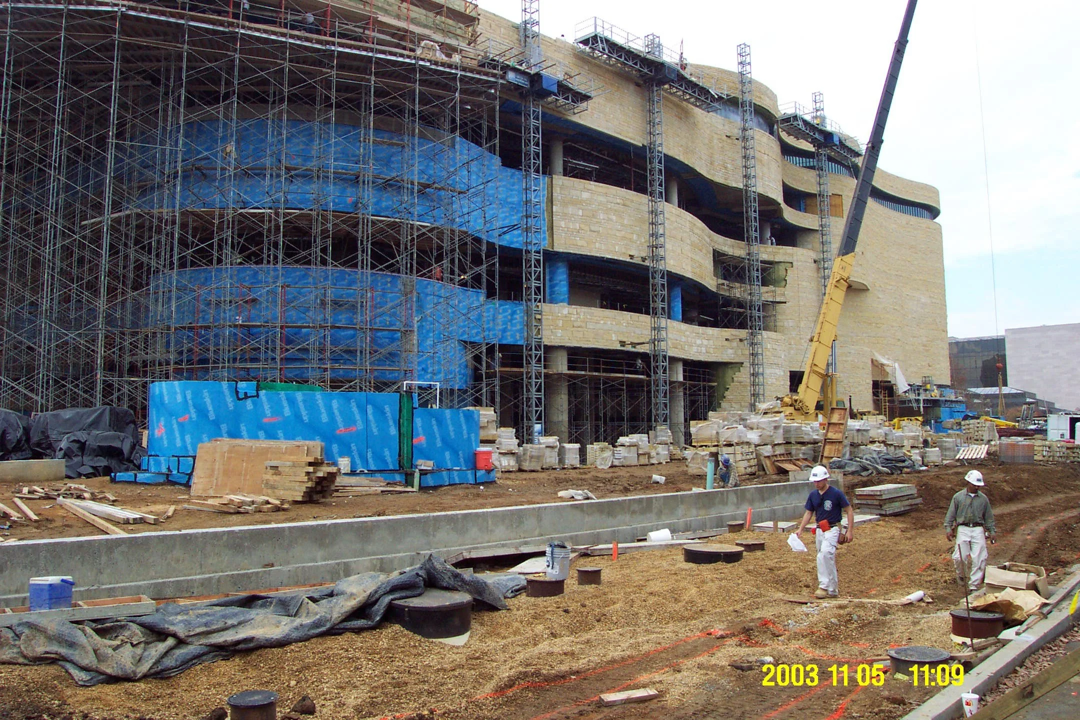 Construction site with workers and scaffolding building a multi-story structure with curved walls. The workers are wearing helmets and safety gear, and there is construction equipment, materials, and a crane present.