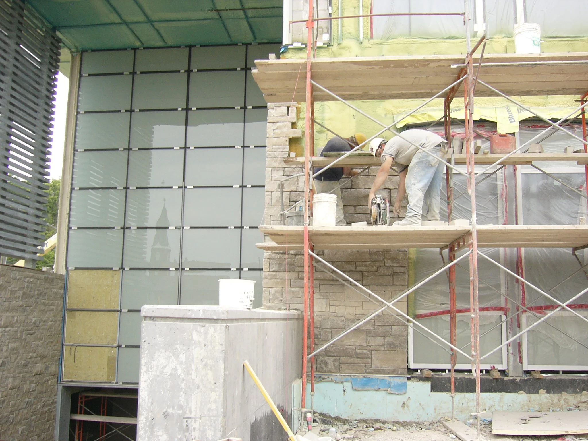 Construction workers on scaffolding installing brick veneer on a building exterior, with reflective glass windows on the left side.