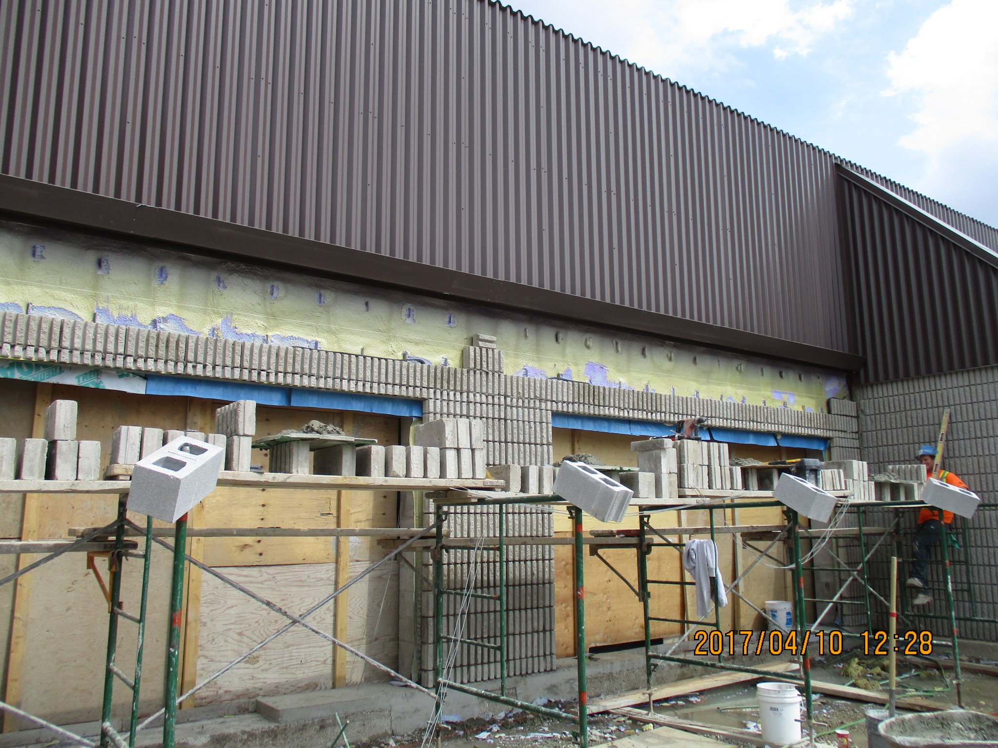 Construction site with workers installing cinder blocks on a building wall, scaffolding in front, and a brown metal exterior wall above, under a partly cloudy sky.