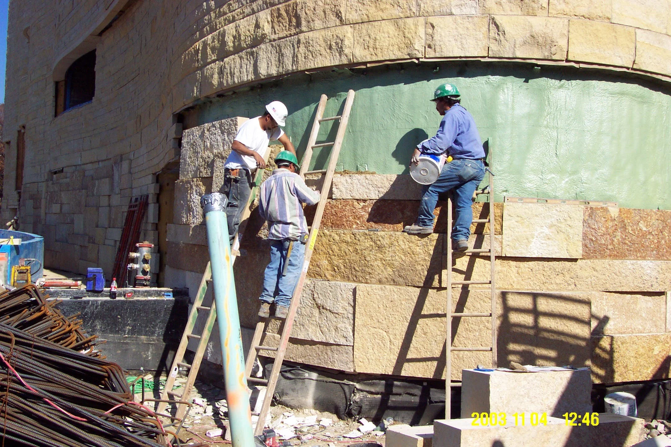 Construction workers wearing safety helmets working on a building façade with large stone blocks and painted sections, using ladders and various tools.