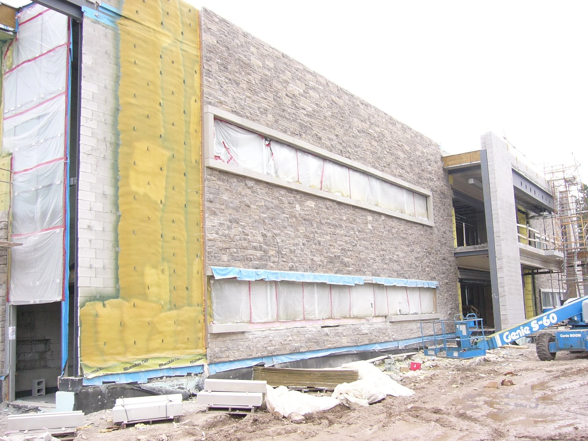 Construction site of a modern building with stone facade, large horizontal windows covered with protective plastic, and scaffolding on the right. Equipment and construction materials are scattered on the ground.