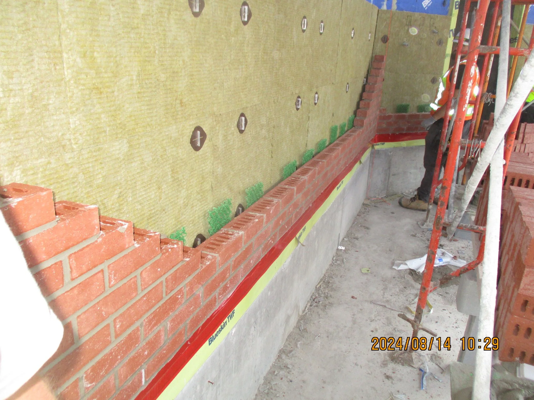 Construction worker building a brick wall on a concrete foundation with scaffolding nearby at a construction site.