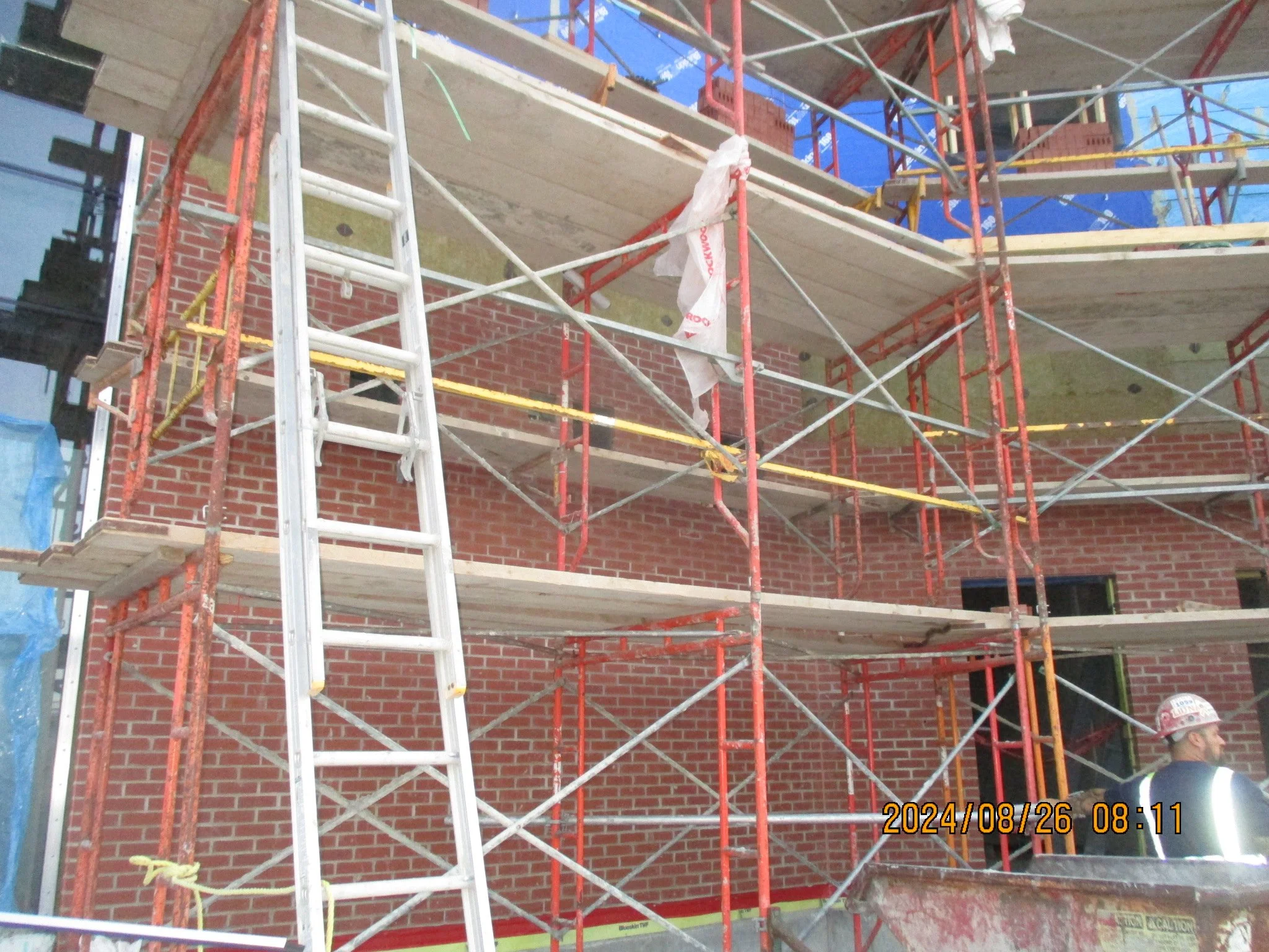 Construction site with red brick building, scaffolding, ladders, and worker wearing safety helmet and vest.