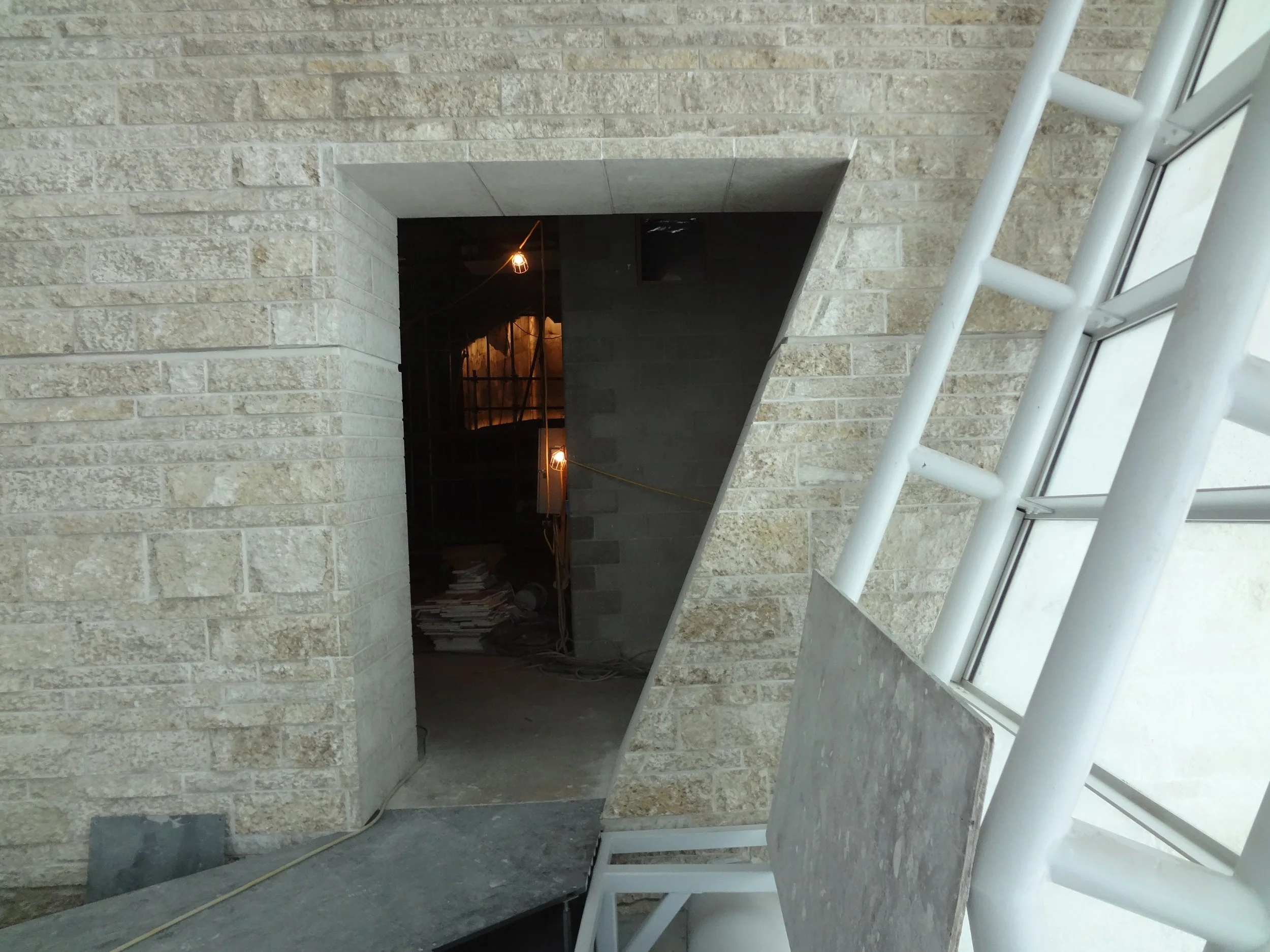 Construction site hallway with brick walls, unfinished flooring, and exposed lights, seen through a window with a white support structure and a piece of construction material leaning against it.