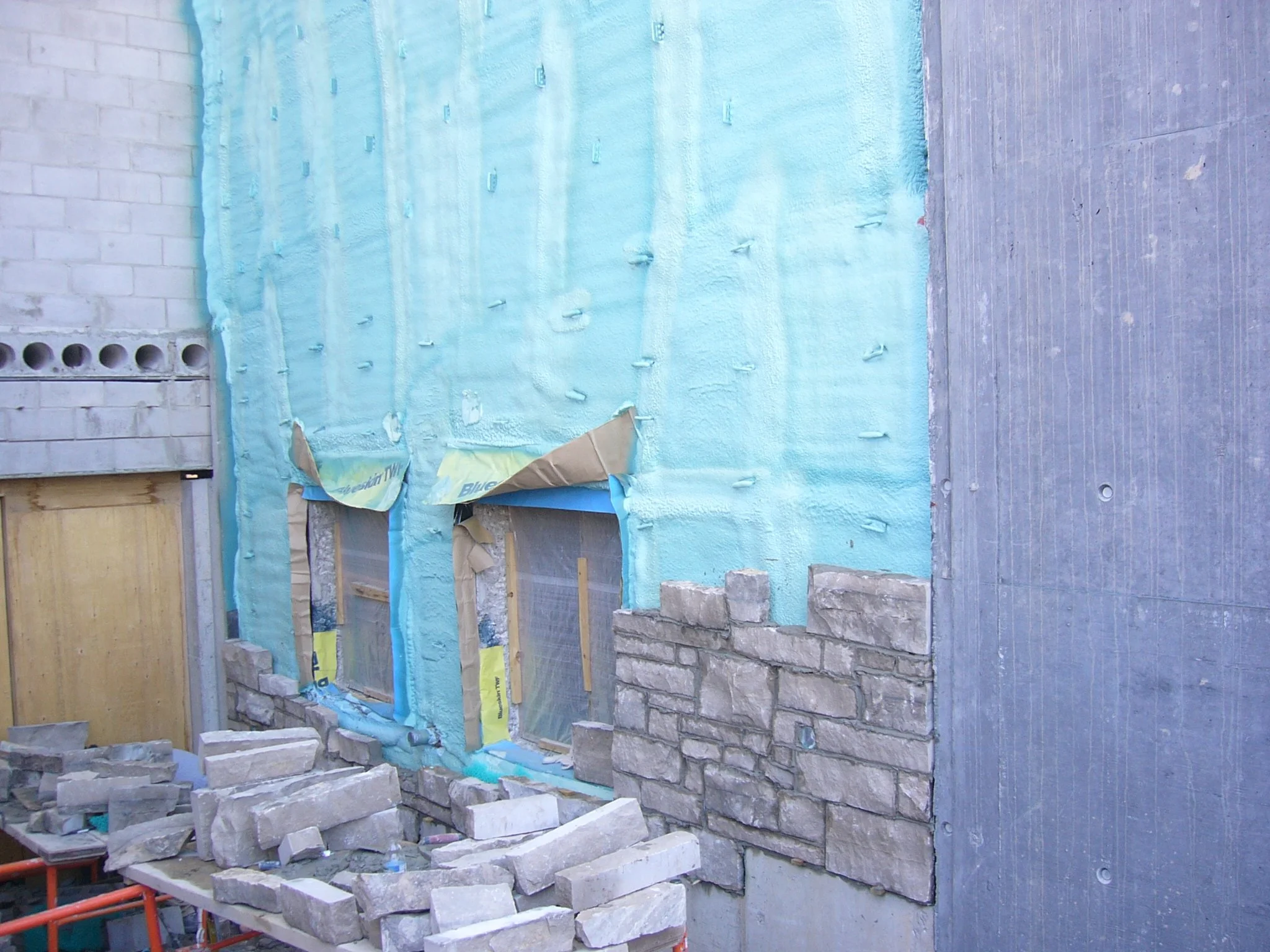 Construction site showing a partially finished wall with insulation foam and stone bricks, with construction materials and tools on the ground.