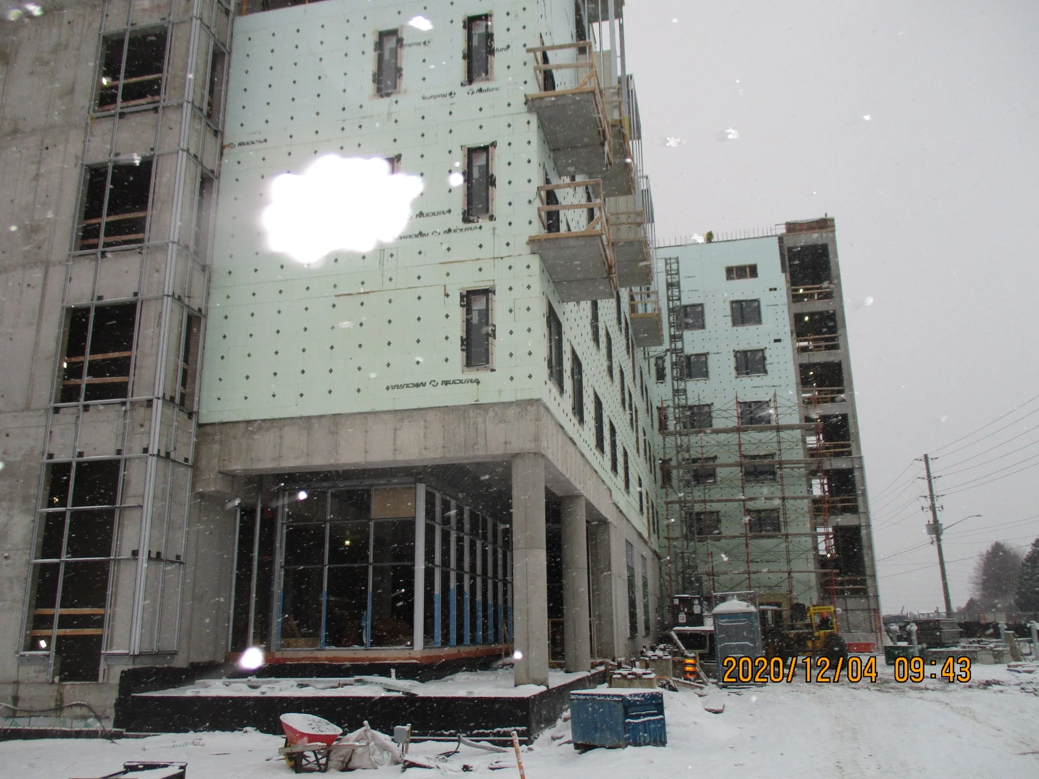 Construction site with multi-story building under construction, scaffolding, snow on ground, and overcast sky.
