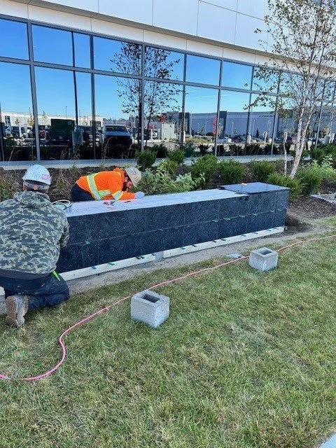Workers installing a large dark gray planter box outside a commercial building with reflective glass windows.