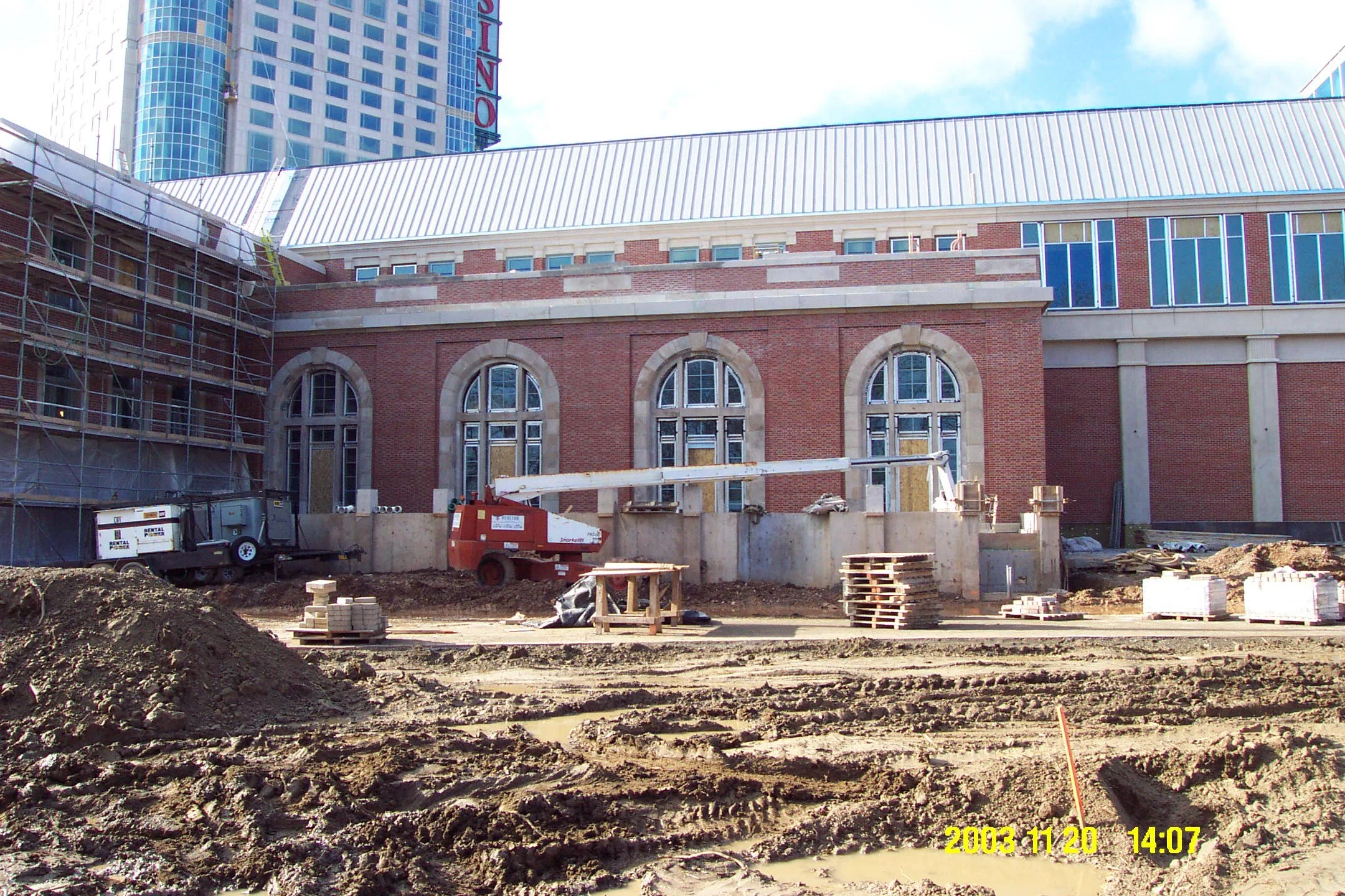 Construction site with muddy ground, construction equipment, stacked pallets, and scaffolding on a brick building with arched windows, in front of modern high-rise buildings.