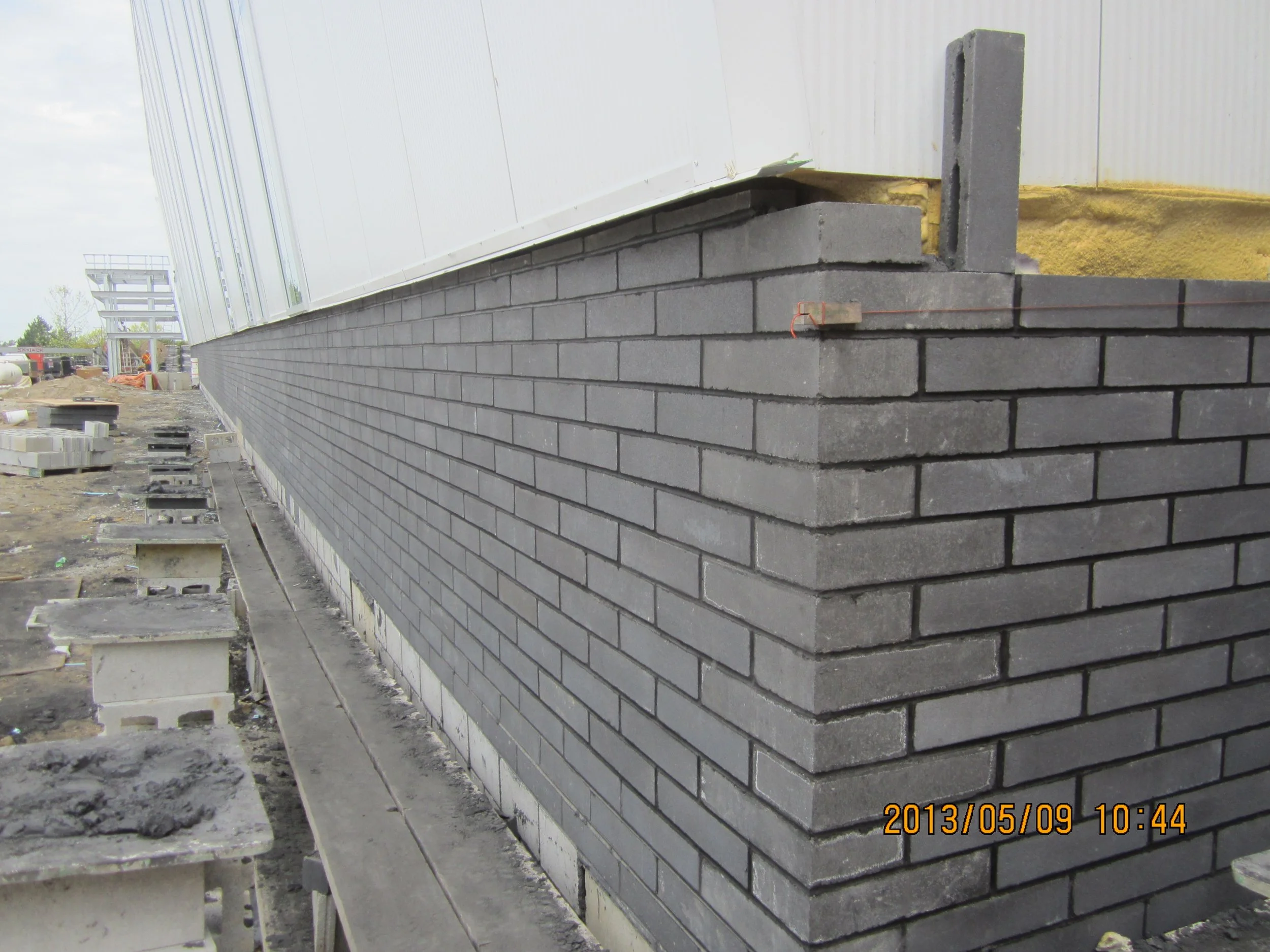 Construction site showing a brick wall with a cinder block foundation and a white exterior wall above. Building materials and tools are scattered nearby.