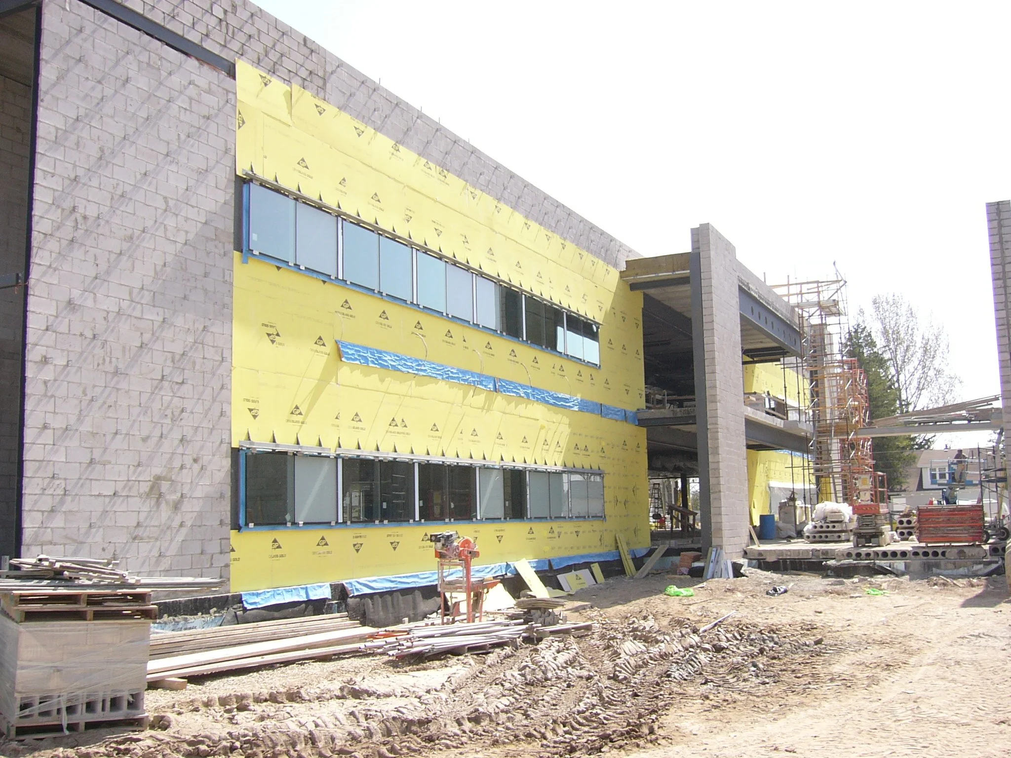 Construction site of a multi-story building with gray brick walls and large windows, partially covered with yellow insulation material, with scaffolding and construction equipment around.