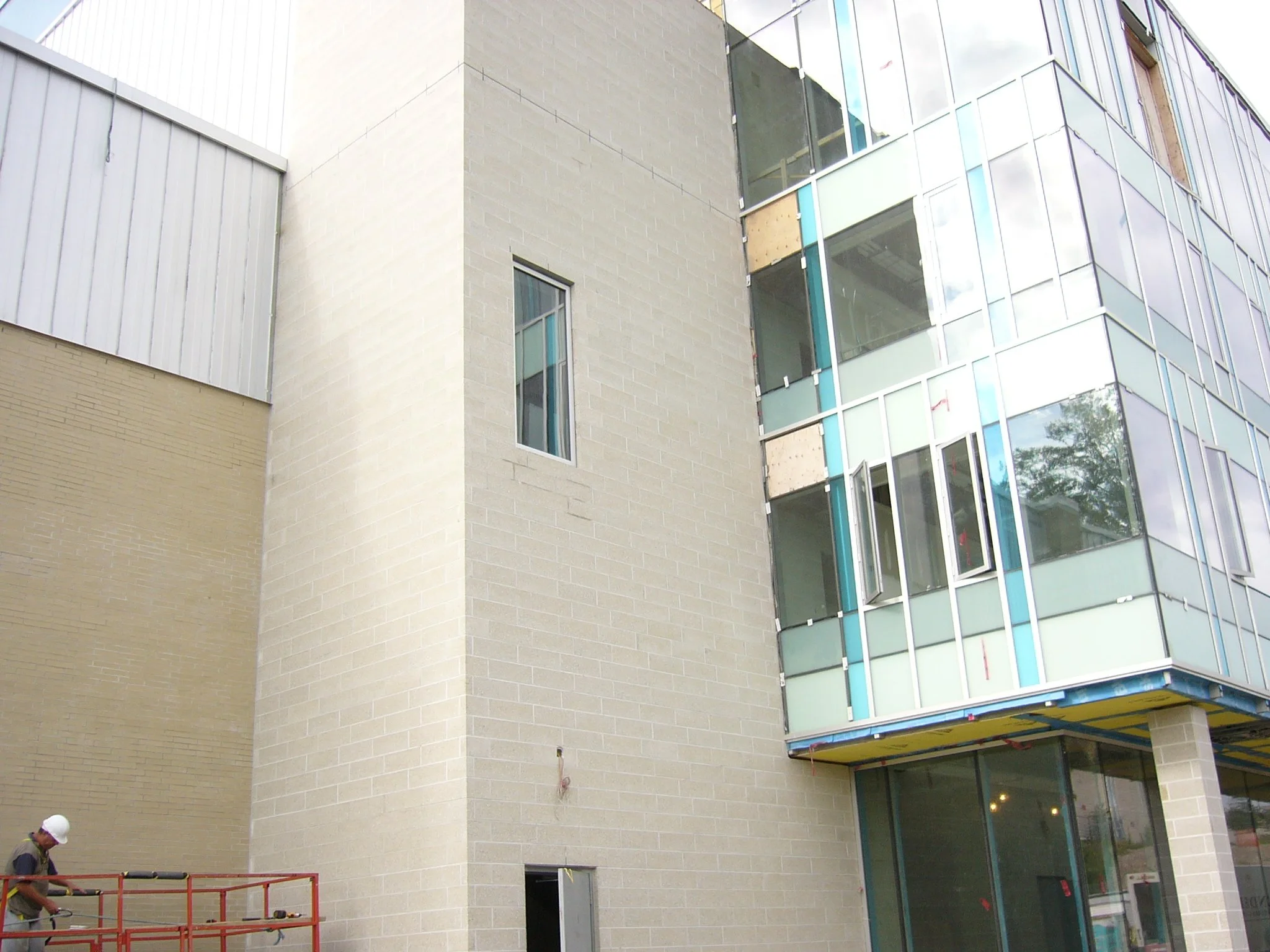 Construction worker wearing a hard hat and safety vest working on building exterior with glass windows and beige brick walls.