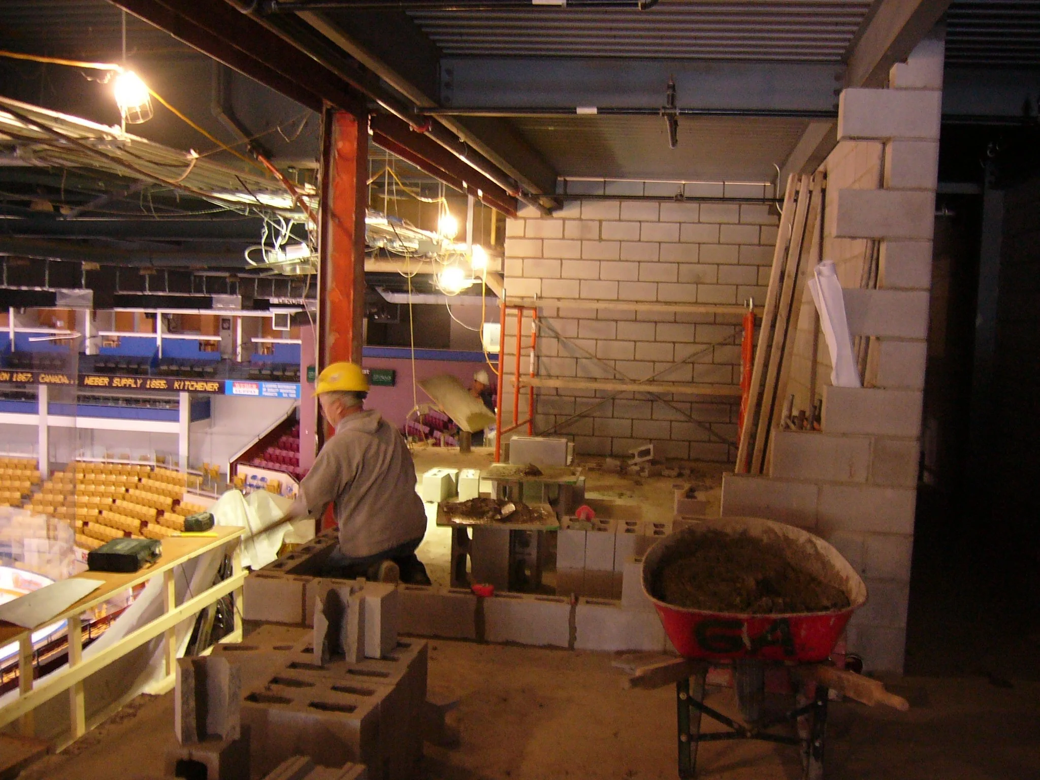Construction workers building a wall inside a stadium, with construction materials and tools around, and a roller wheelbarrow filled with dirt in the foreground.