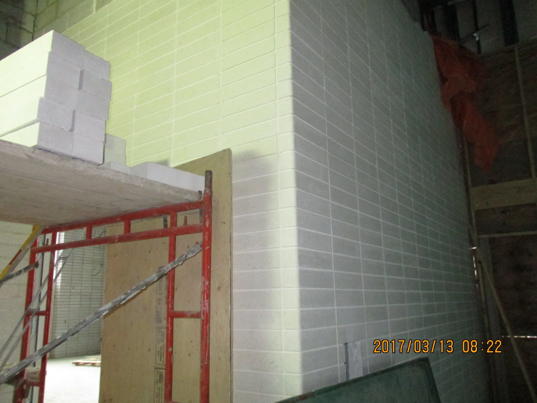 Construction site showing a corner of a building with tiled walls, a scaffolding with red metal supports, and some construction materials such as a stack of bricks and a wooden board. There is an orange tarp visible in the background under a wooden r