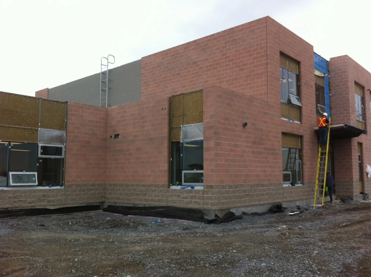 Construction workers building a multi-story brick building with large windows, ladder, and scaffolding, on a dirt lot.
