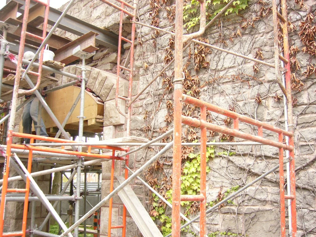 Construction scaffold set up next to a stone wall with ivy plants, with a worker partially visible on the left side.