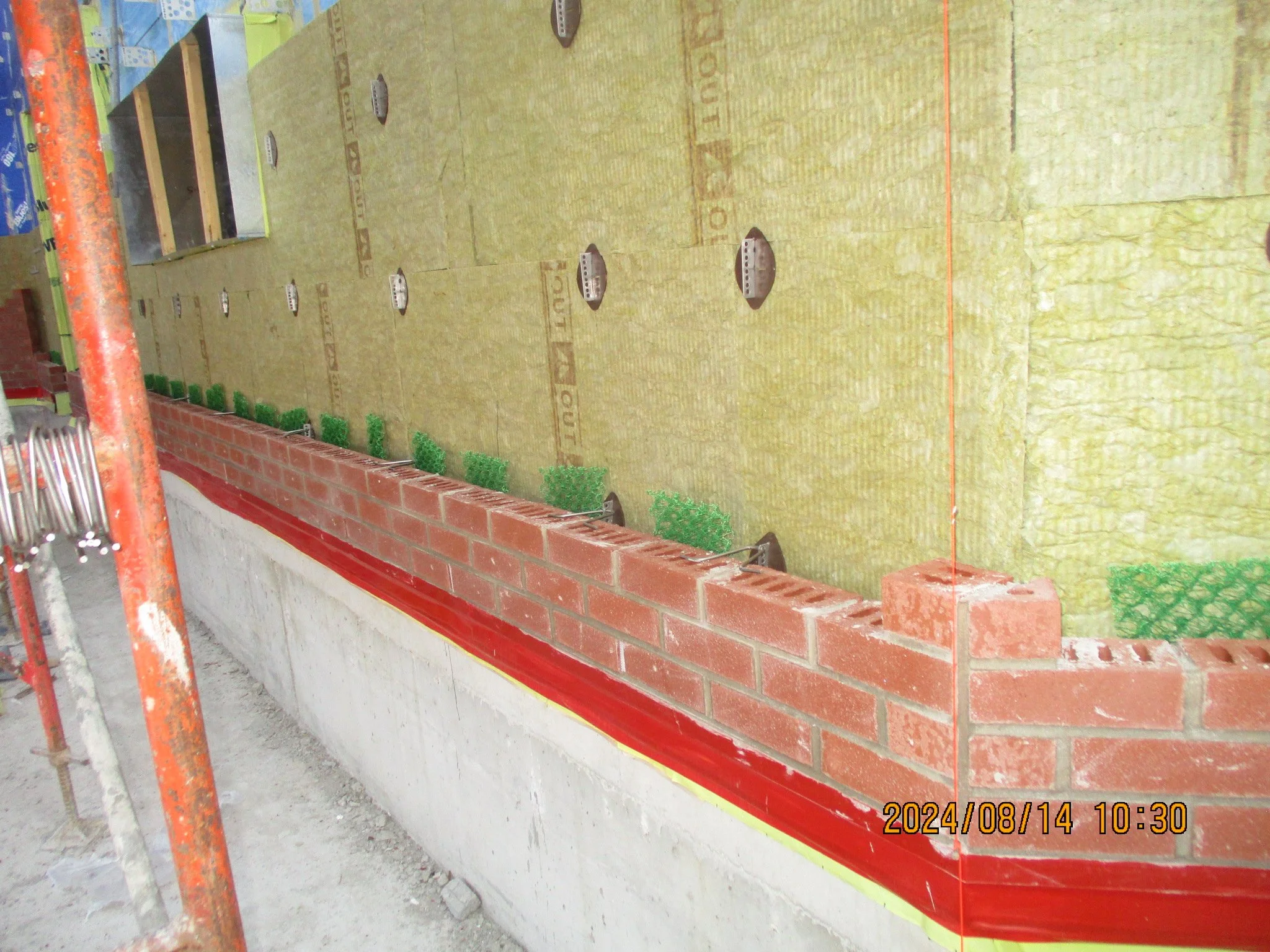 Construction site with a partially built brick wall along a concrete foundation. Insulation material is visible inside the wall, and there are small green bushes lined up at the base of the wall. Construction equipment and tools are visible on the si