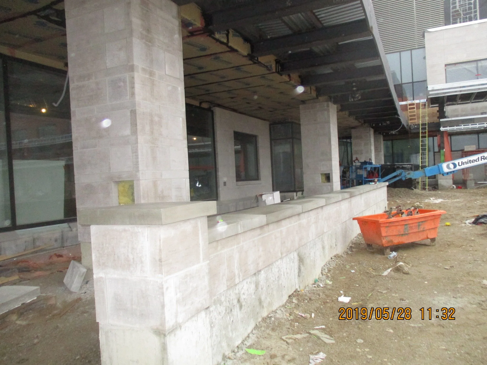 Construction site with a building under construction, scaffolding, and construction equipment, including a blue lift and orange container, and dirt ground scattered with debris.