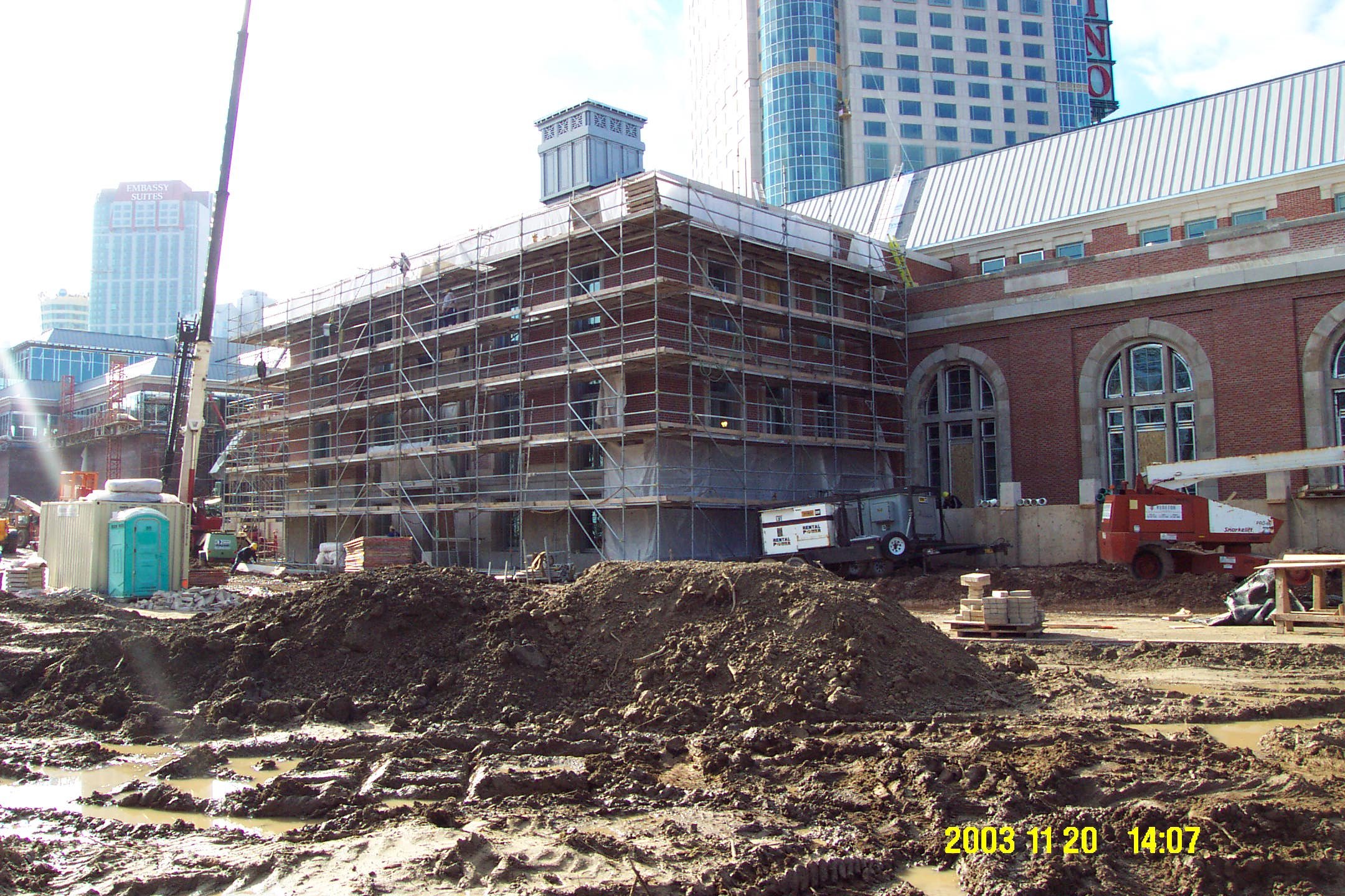 Construction site with a partially built brick building surrounded by scaffolding, construction equipment, and muddy ground in an urban area with tall buildings in the background.