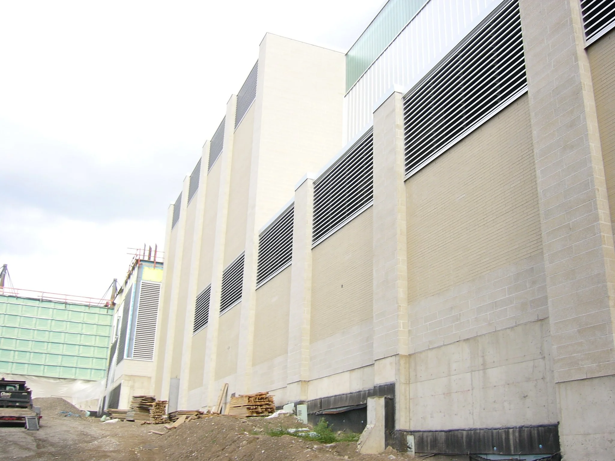 Modern building under construction with beige brick walls, metal ventilation grates, and construction materials and dirt in the foreground.